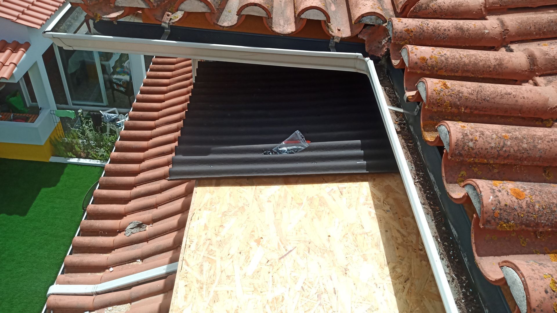 Reparo de telhado, detalhe. Telhas danificadas, compensado novo, material ondulado preto, calha, gramado verde ao fundo.