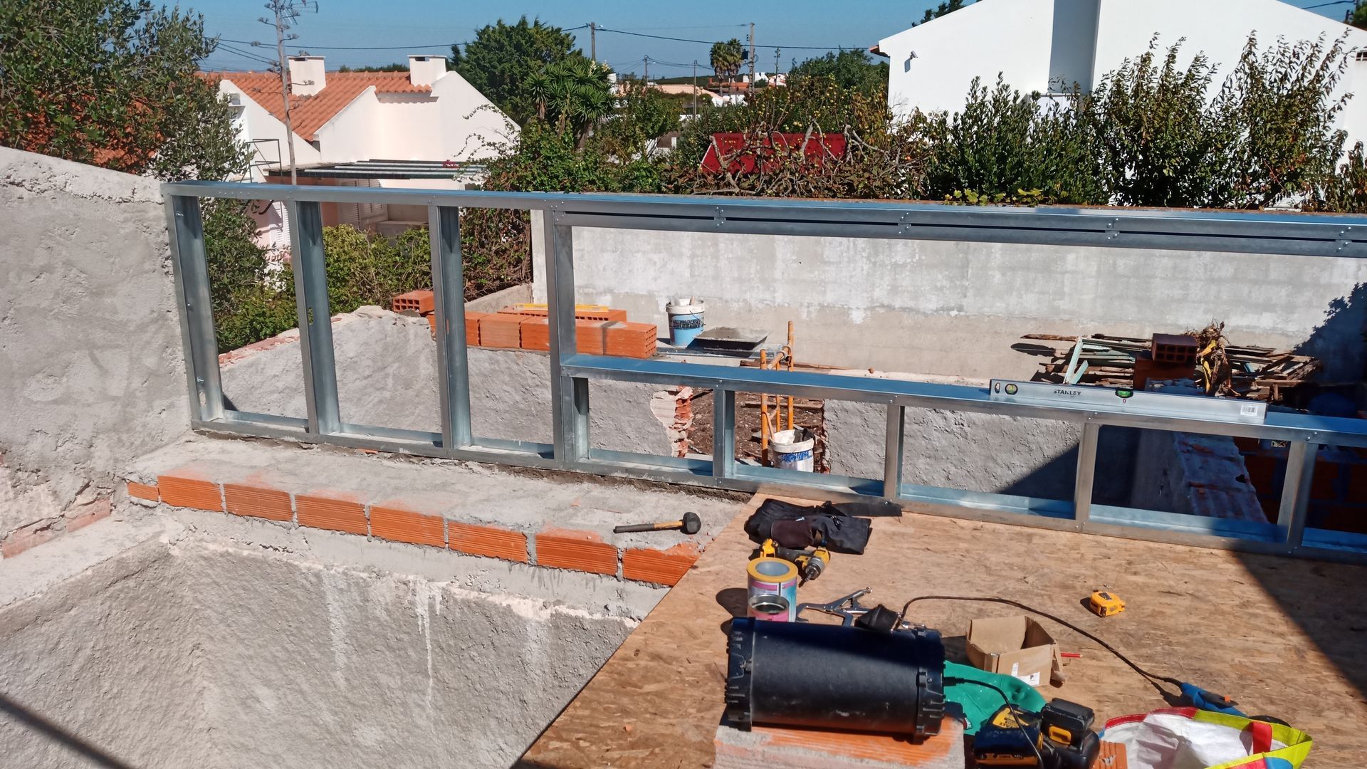Canteiro de obras com estrutura metálica sobre base de tijolos, ferramentas e materiais presentes. Ao ar livre, ensolarado.
