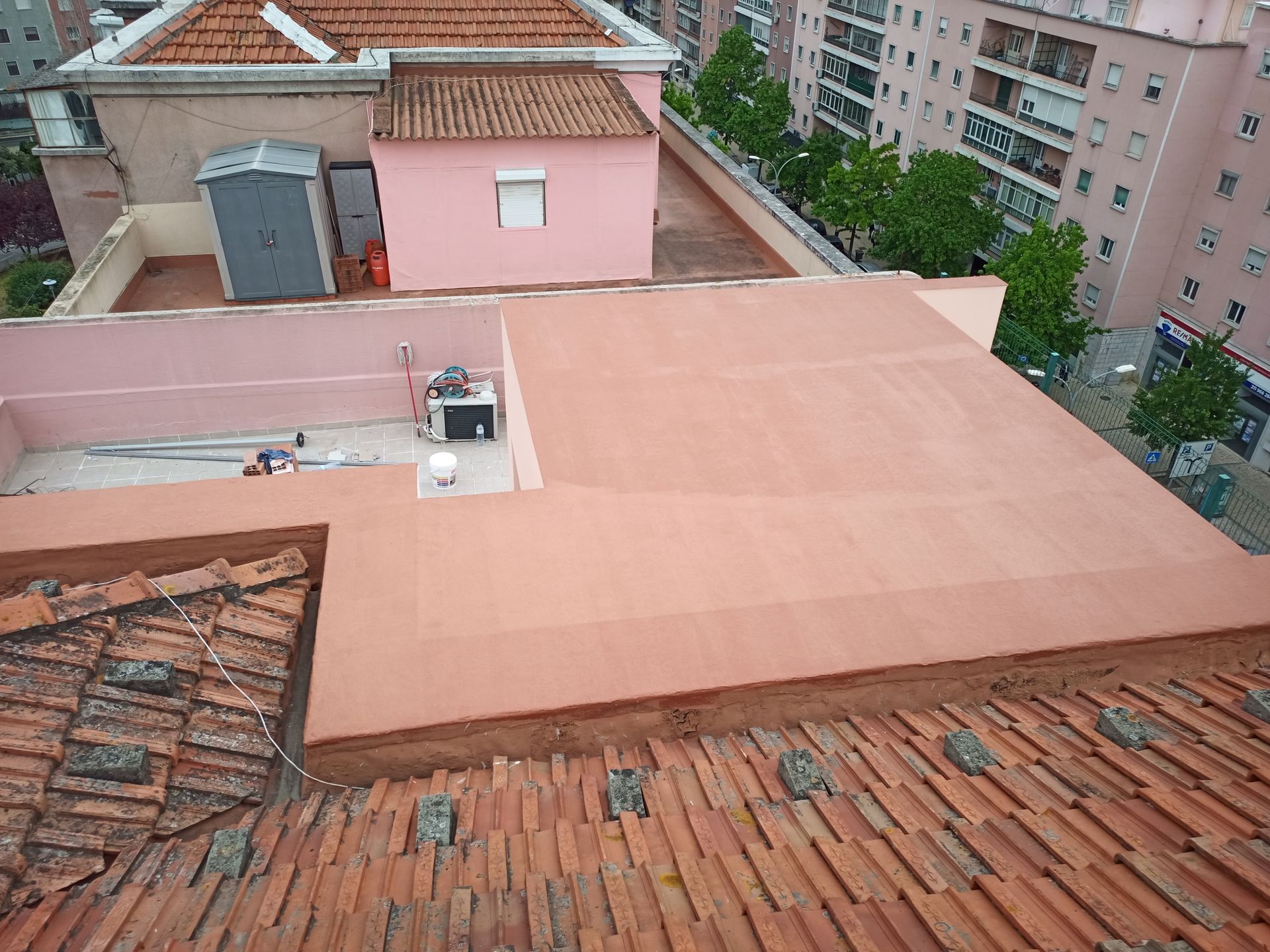 Vista de um terraço: telhado de telha vermelha em primeiro plano, telhado plano vermelho com equipamentos e prédio rosa.
