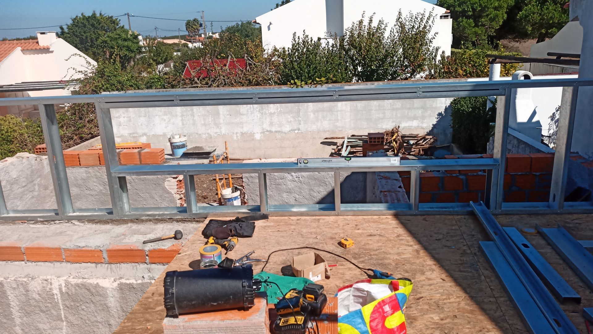 Canteiro de obras: estrutura metálica e alvenaria em um telhado, ferramentas e materiais espalhados. Ambiente externo ensolarado.