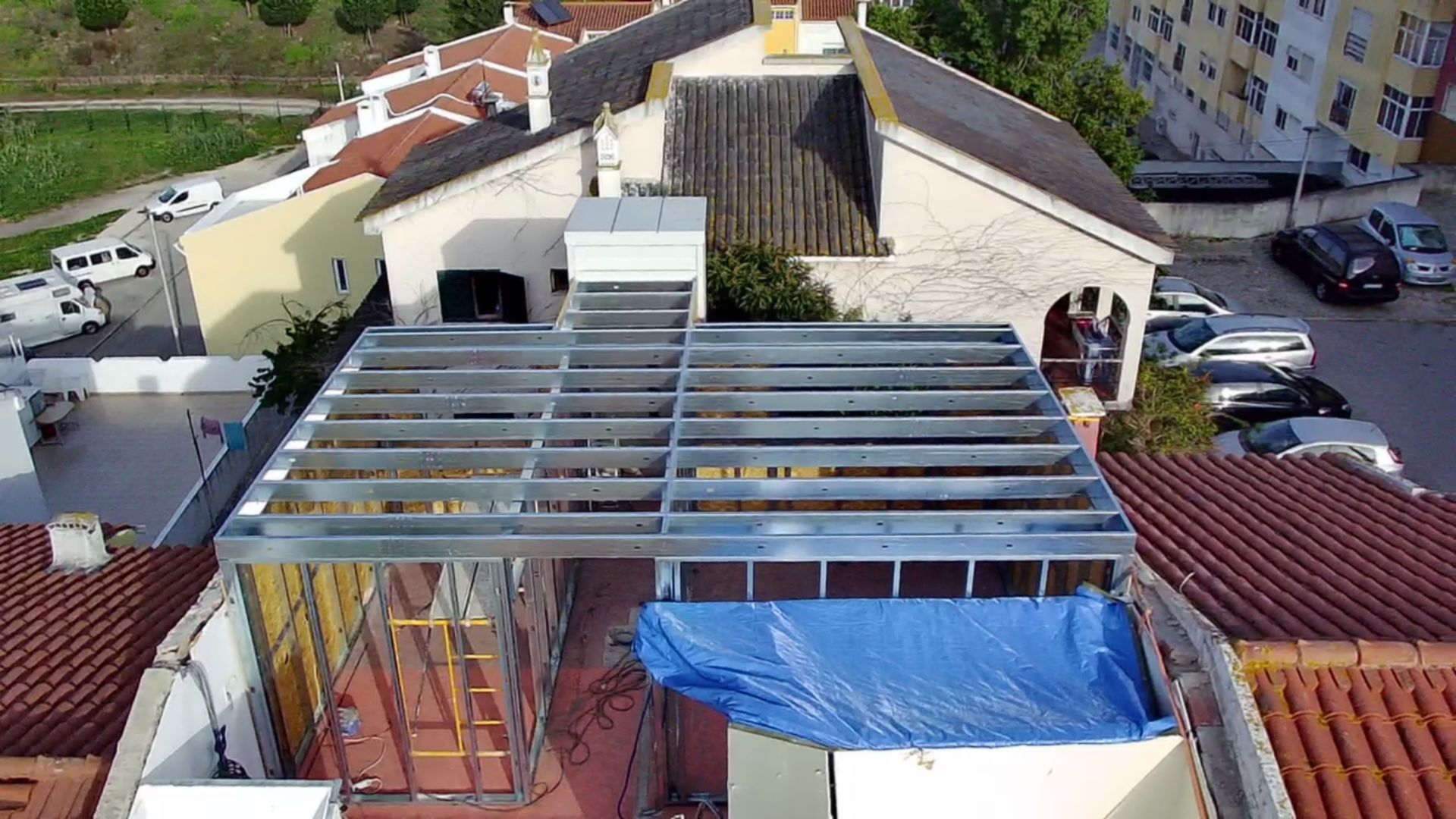 Construção de uma estrutura metálica no telhado de um edifício, cercada por outros telhados e carros estacionados.