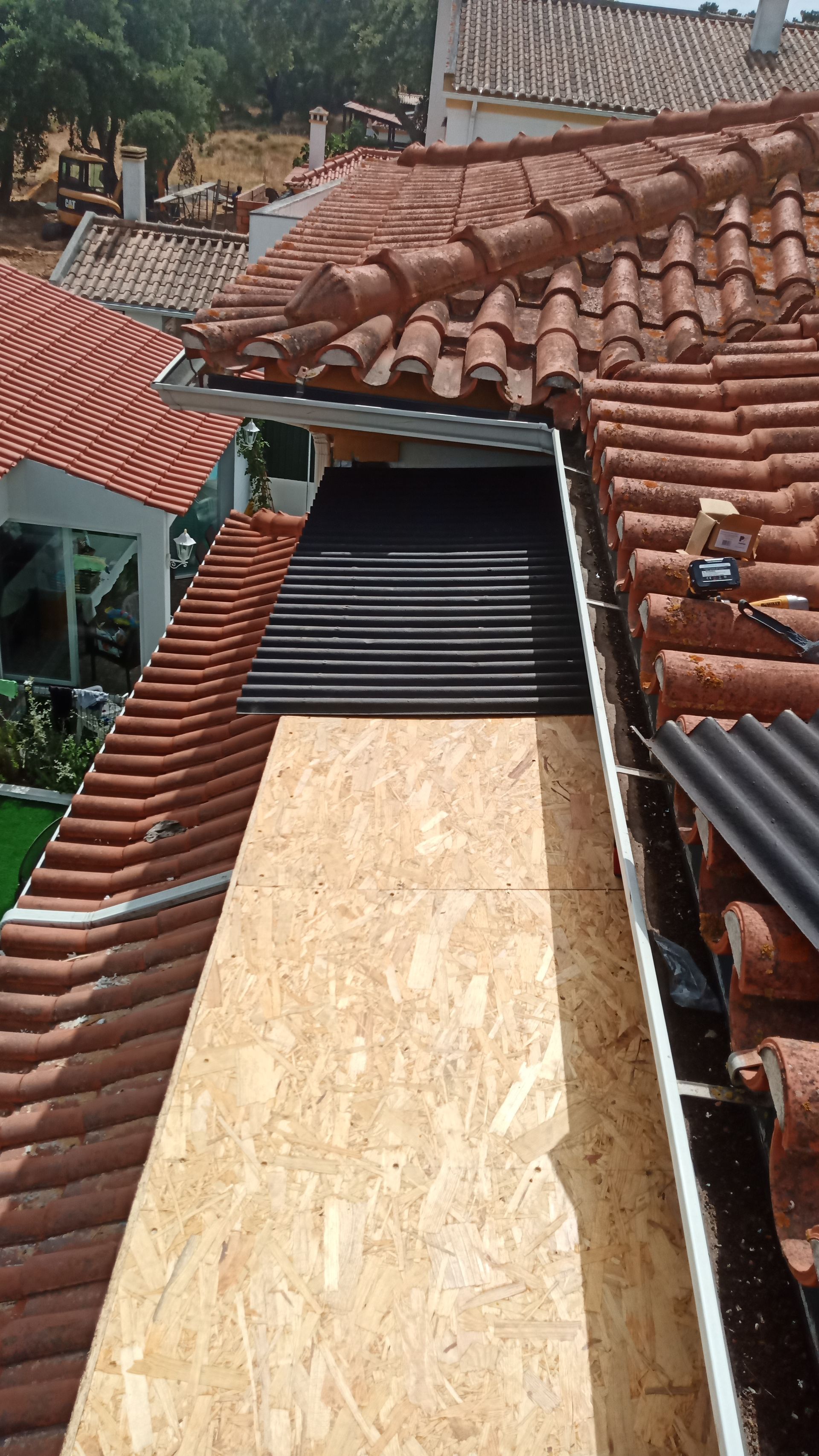 Cena em um telhado com telhas laranja, madeira compensada e material ondulado preto; calhas e edifícios visíveis.