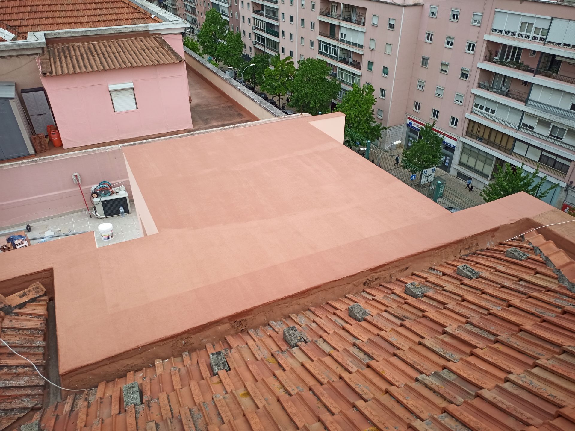 Vista aérea de telhados. Telhado de telha vermelha em primeiro plano, telhado plano rosa atrás com equipamentos e edifícios ao fundo.