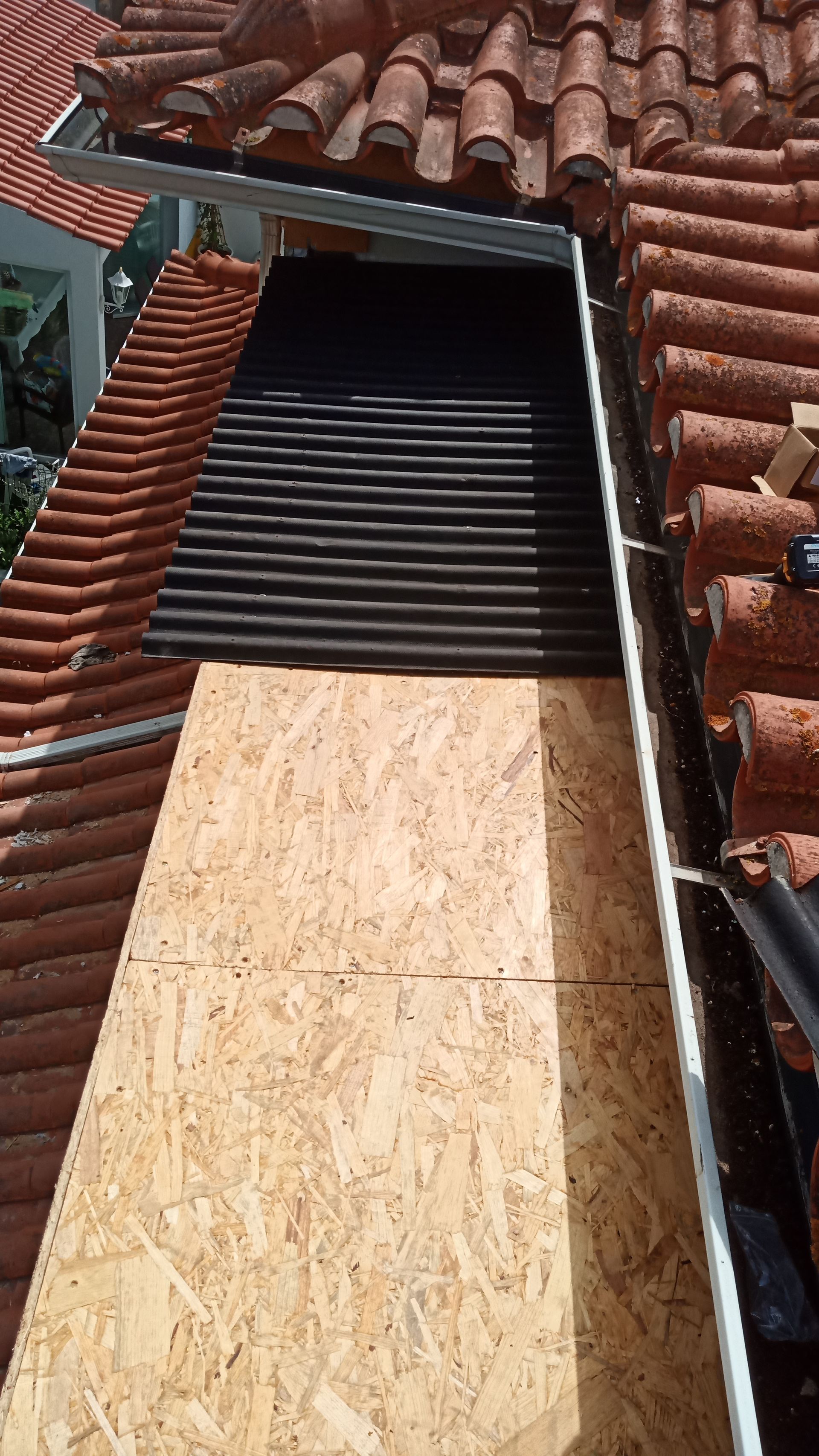 Telhado com telhas laranja, painéis ondulados pretos e uma grande tábua de madeira. Calhas e um vislumbre da casa são visíveis.