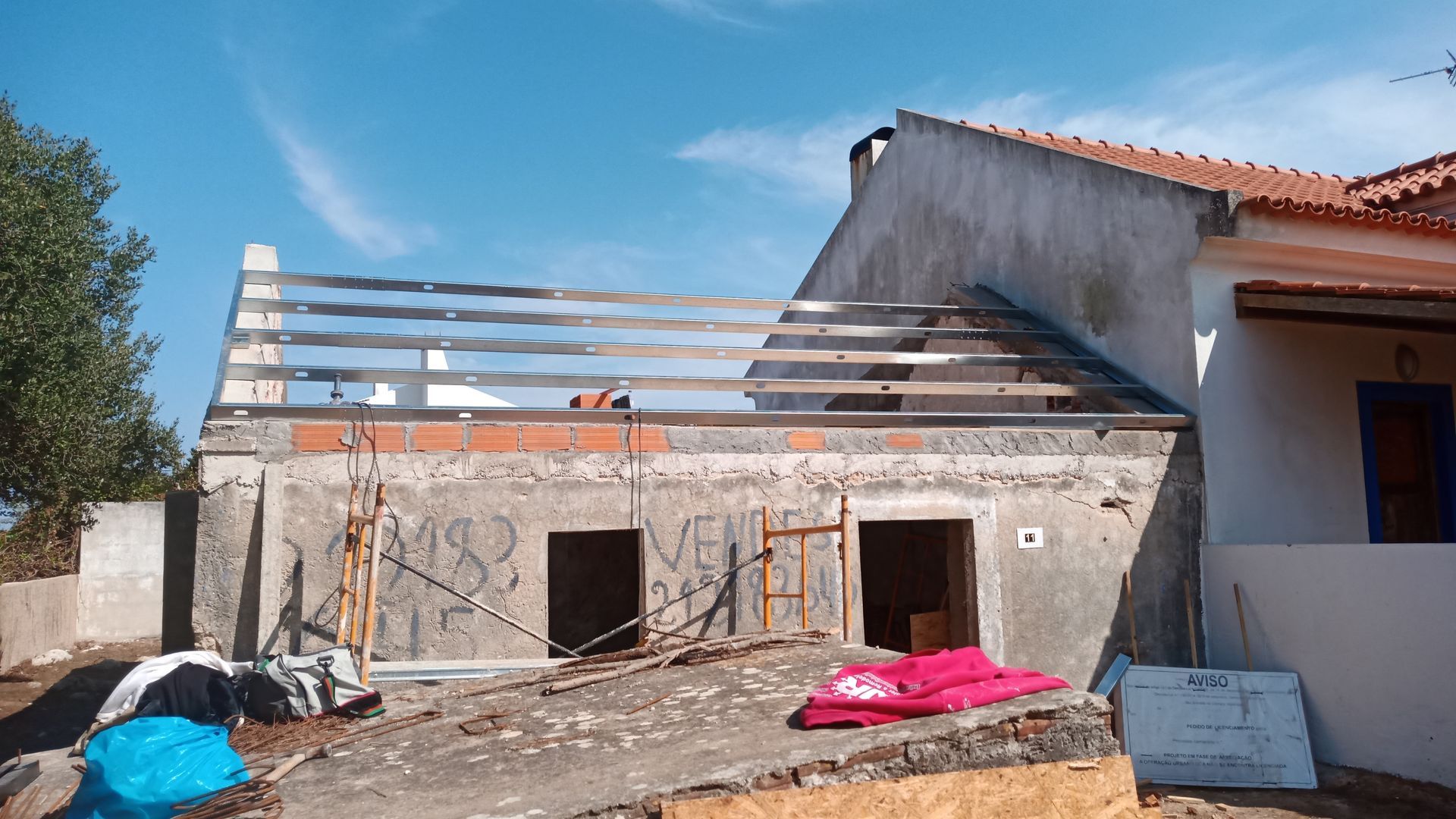 Prédio em construção com vigas expostas, alvenaria e parte do telhado aberto, contra um céu azul.