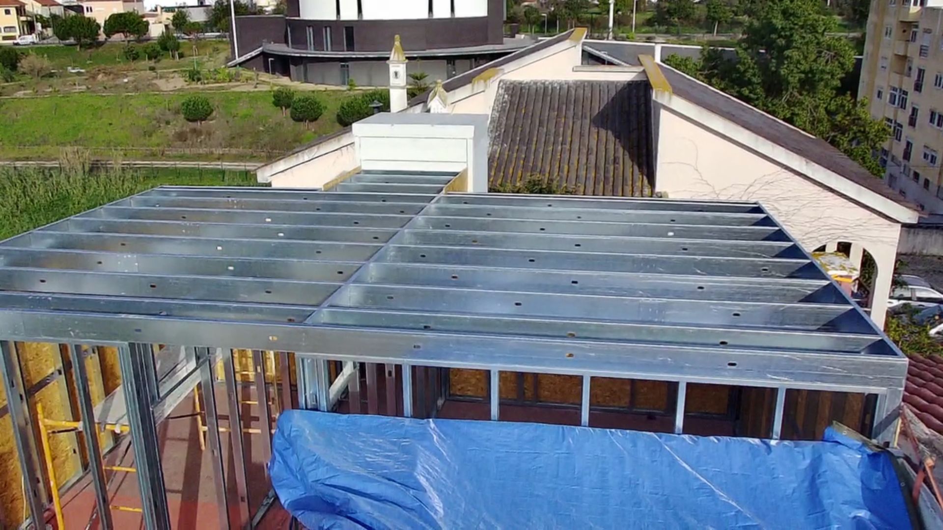 Estrutura metálica do telhado de um edifício em construção, com outros edifícios e vegetação ao redor.