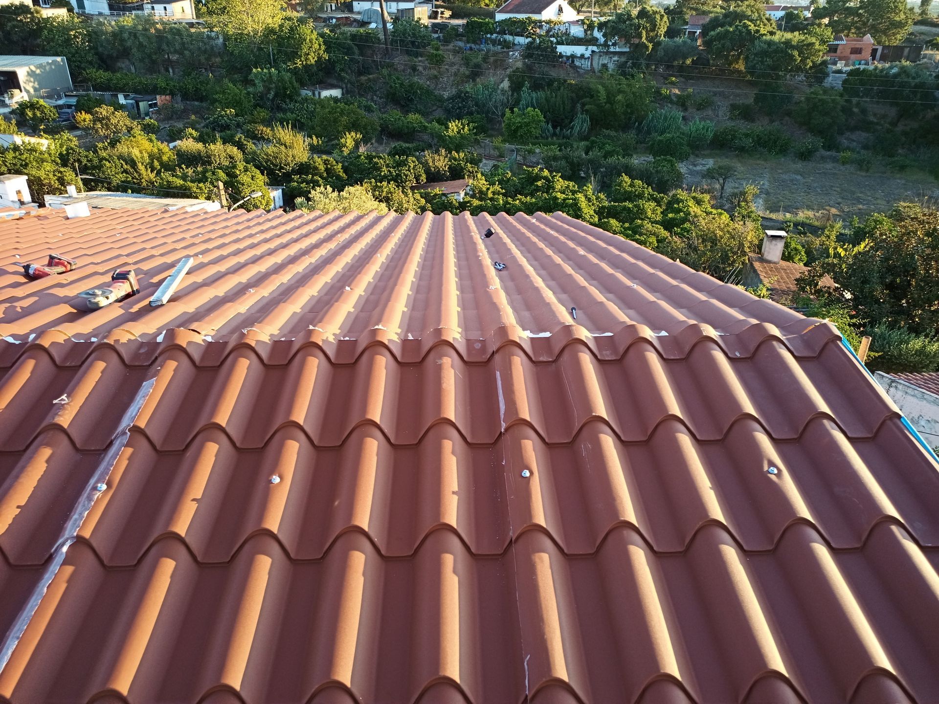 Close-up de um telhado de telhas onduladas marrons, com ferramentas sobre a superfície. Prédios e árvores são visíveis ao fundo.