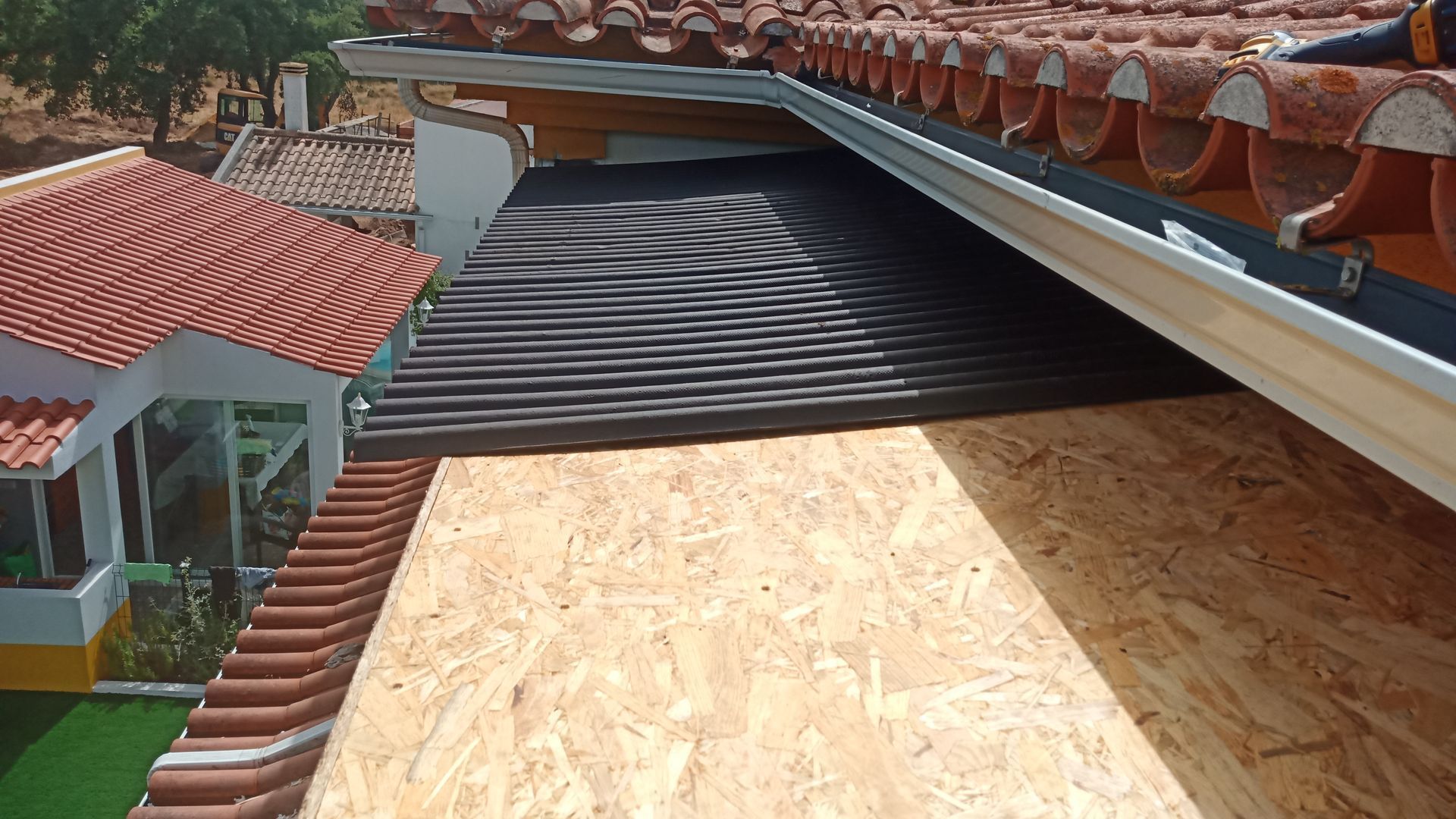 Detalhe de um telhado parcialmente construído com cobertura metálica preta, subcobertura de madeira e telhado de telhas adjacente.