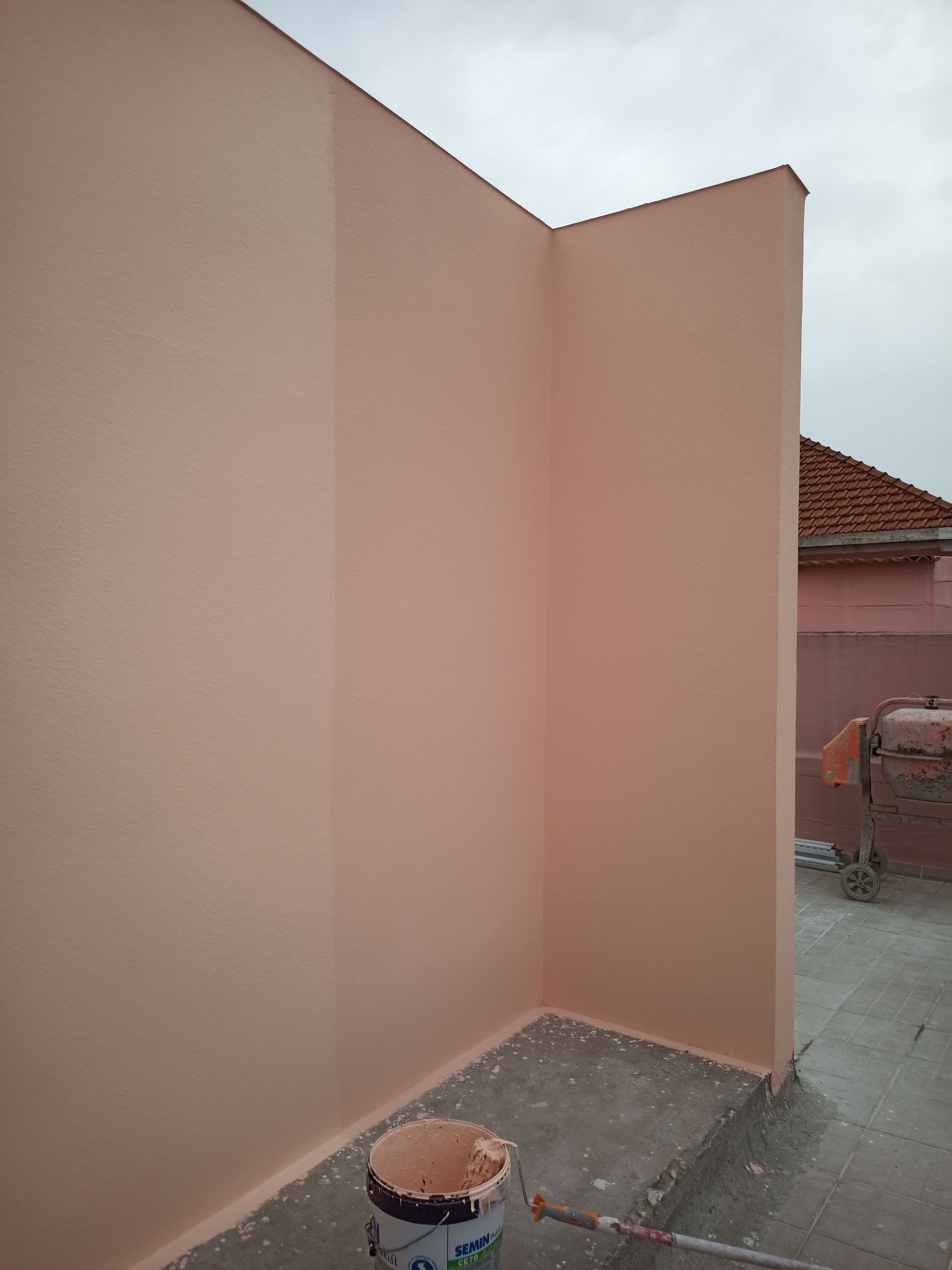 Canto de um prédio pintado de rosa com um balde de tinta no chão de concreto.