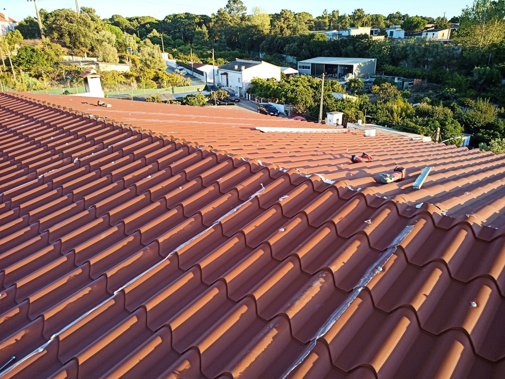 Telhado de telha vermelha com vista para uma área residencial, com árvores verdes ao fundo sob a luz do sol.