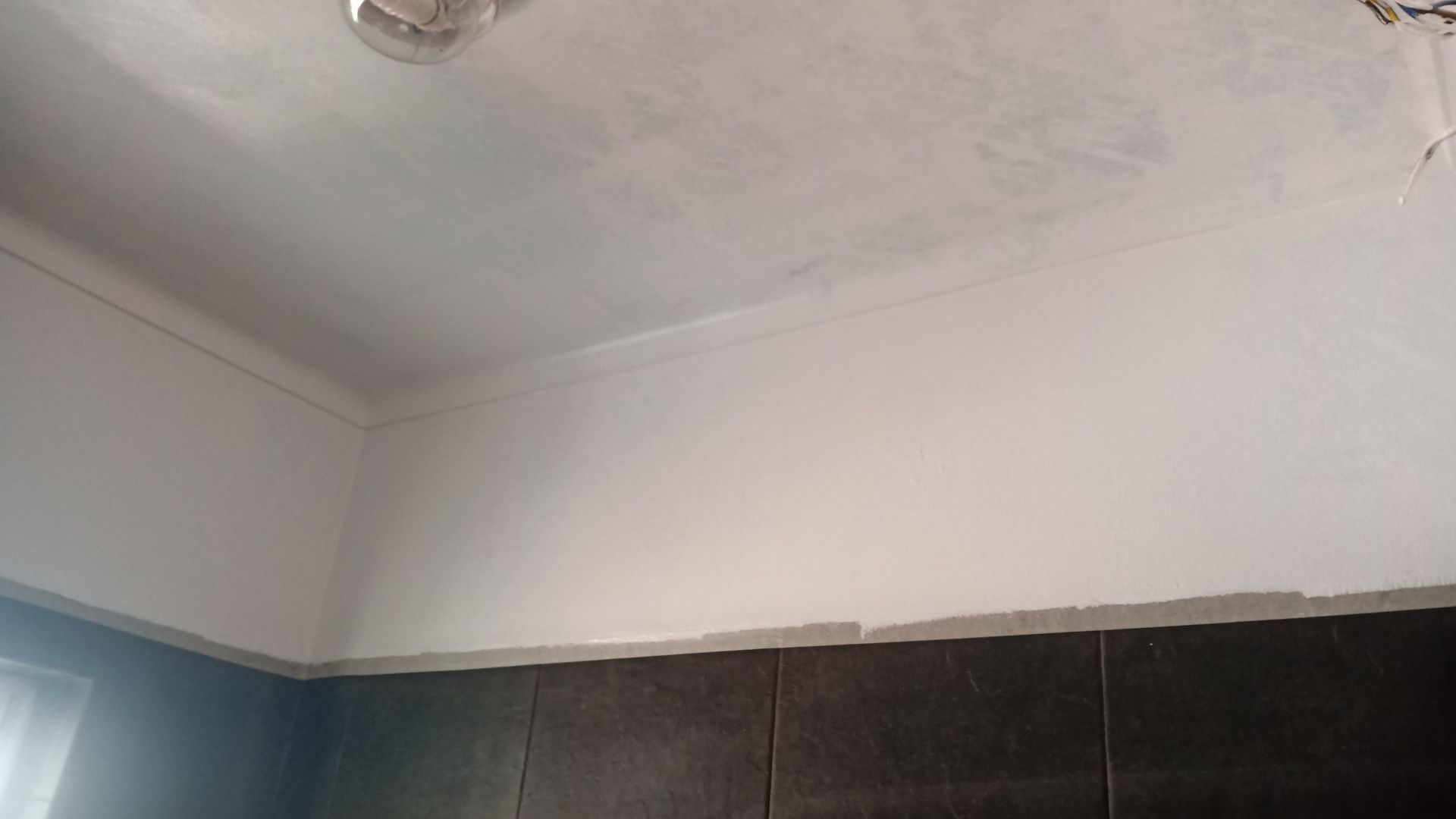 Canto de uma sala com teto e paredes brancas, azulejo escuro. Algumas paredes estão inacabadas.