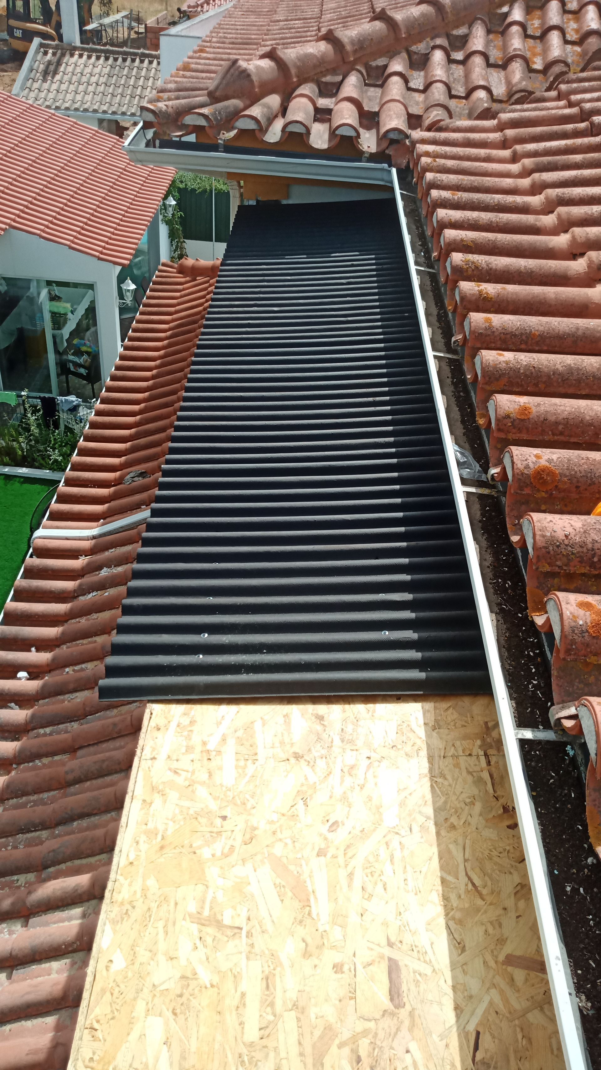 Vista aérea de um telhado em construção. Painéis ondulados pretos entre telhas vermelhas e uma base de madeira compensada.