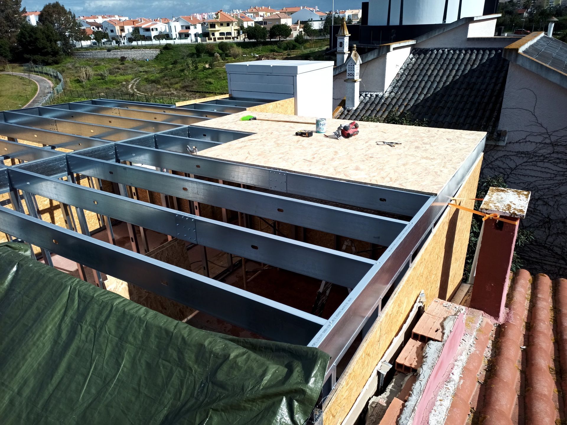 Estrutura metálica de um telhado em construção, com materiais de construção e telhados visíveis ao fundo.
