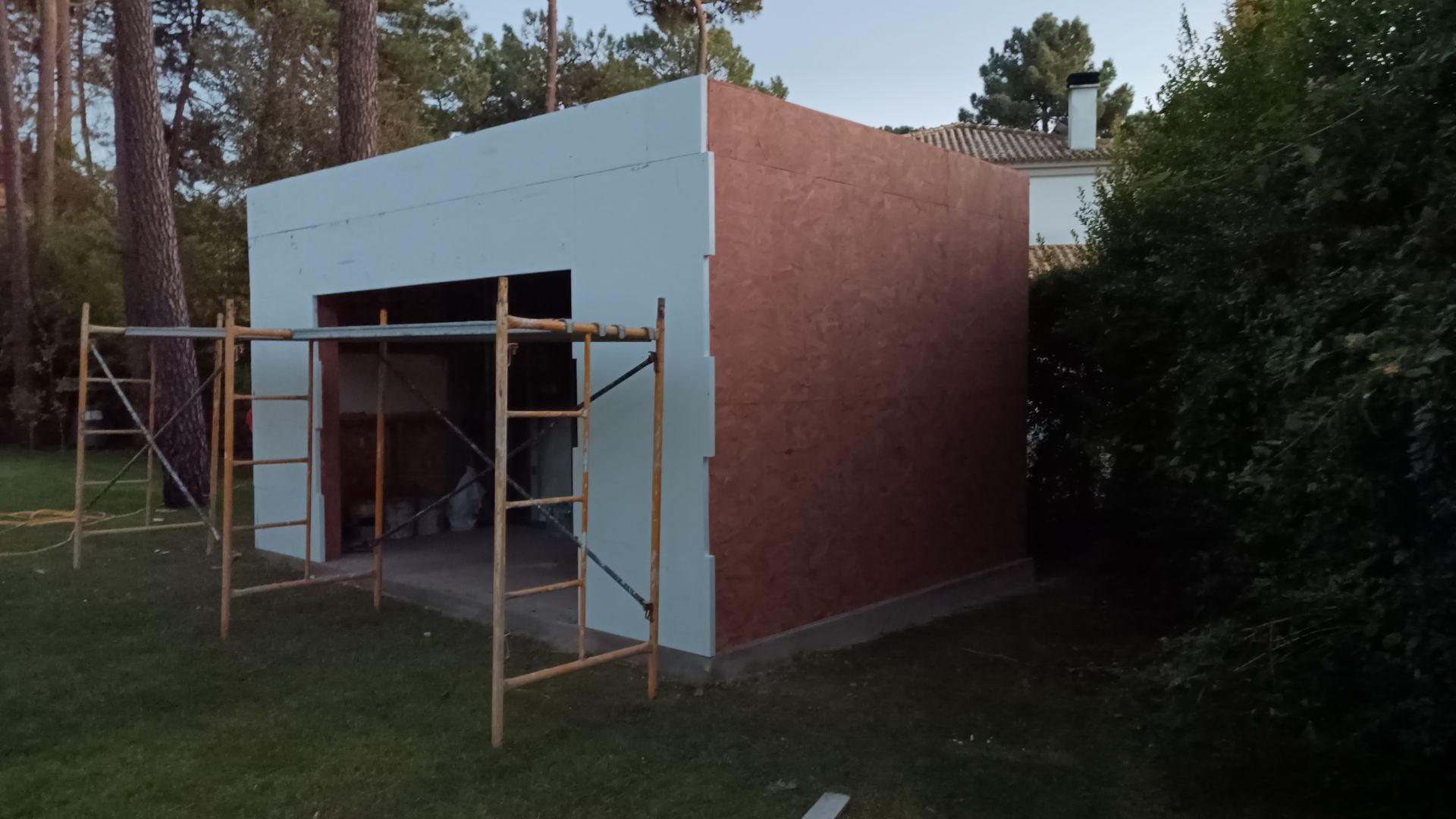 Garagem em construção com paredes brancas e vermelhas, cercada por andaimes e grama.
