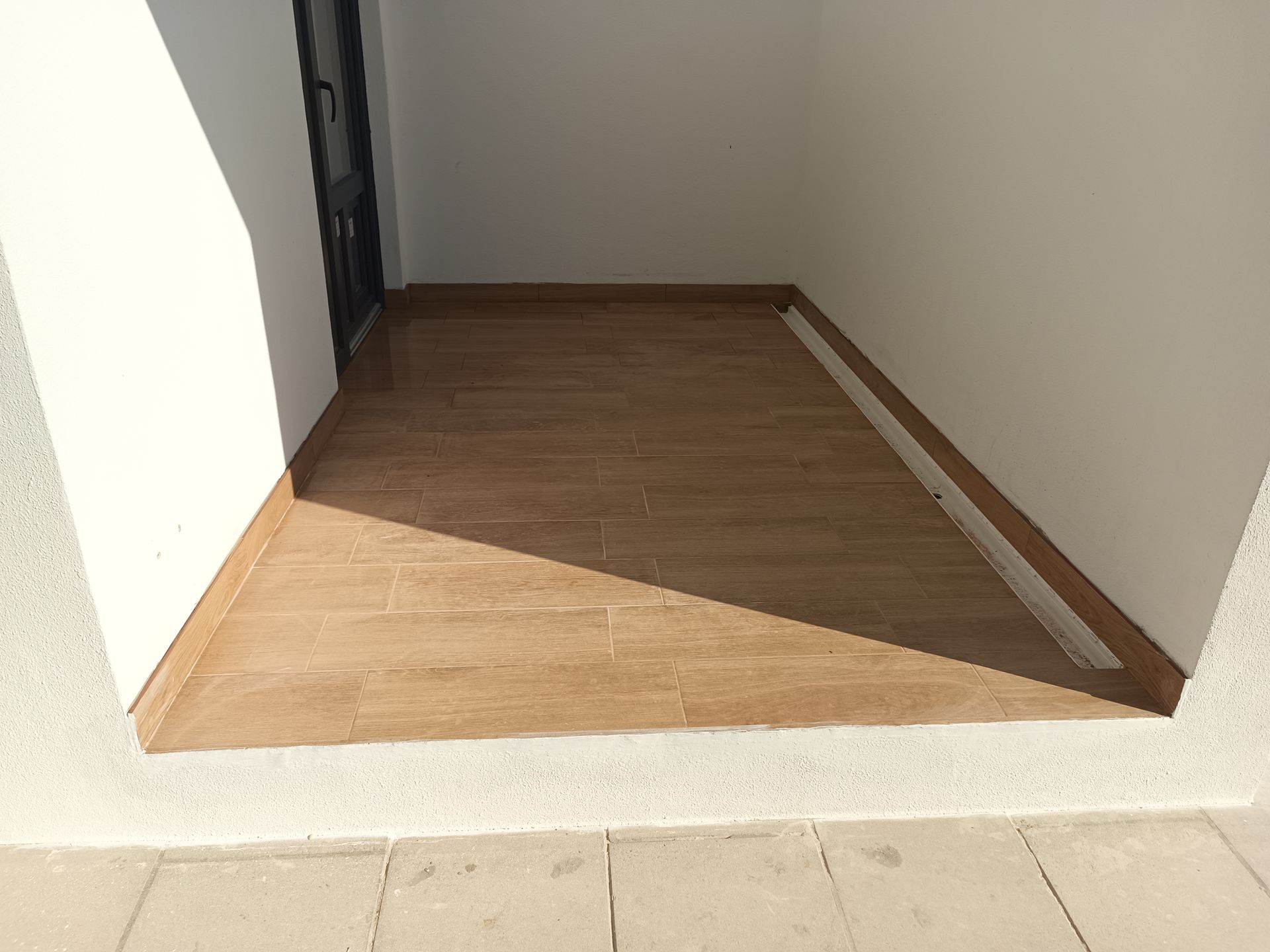 Pequeno pátio coberto com piso de porcelanato imitando madeira. Paredes brancas, porta nos fundos, luz solar e sombra.