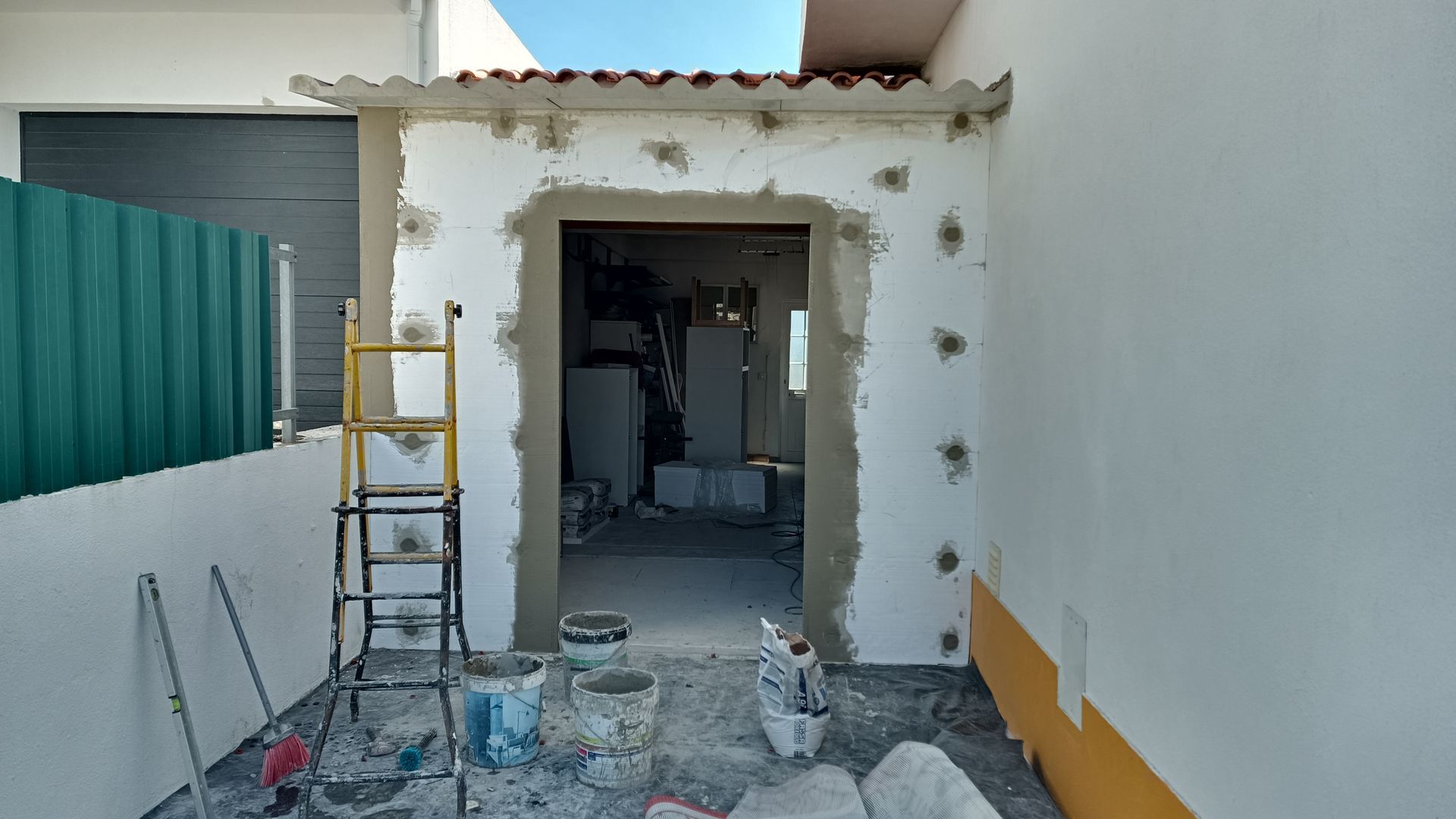 Uma porta em construção, cercada por paredes brancas com argamassa cinza e um pequeno telhado, com uma escada em frente.