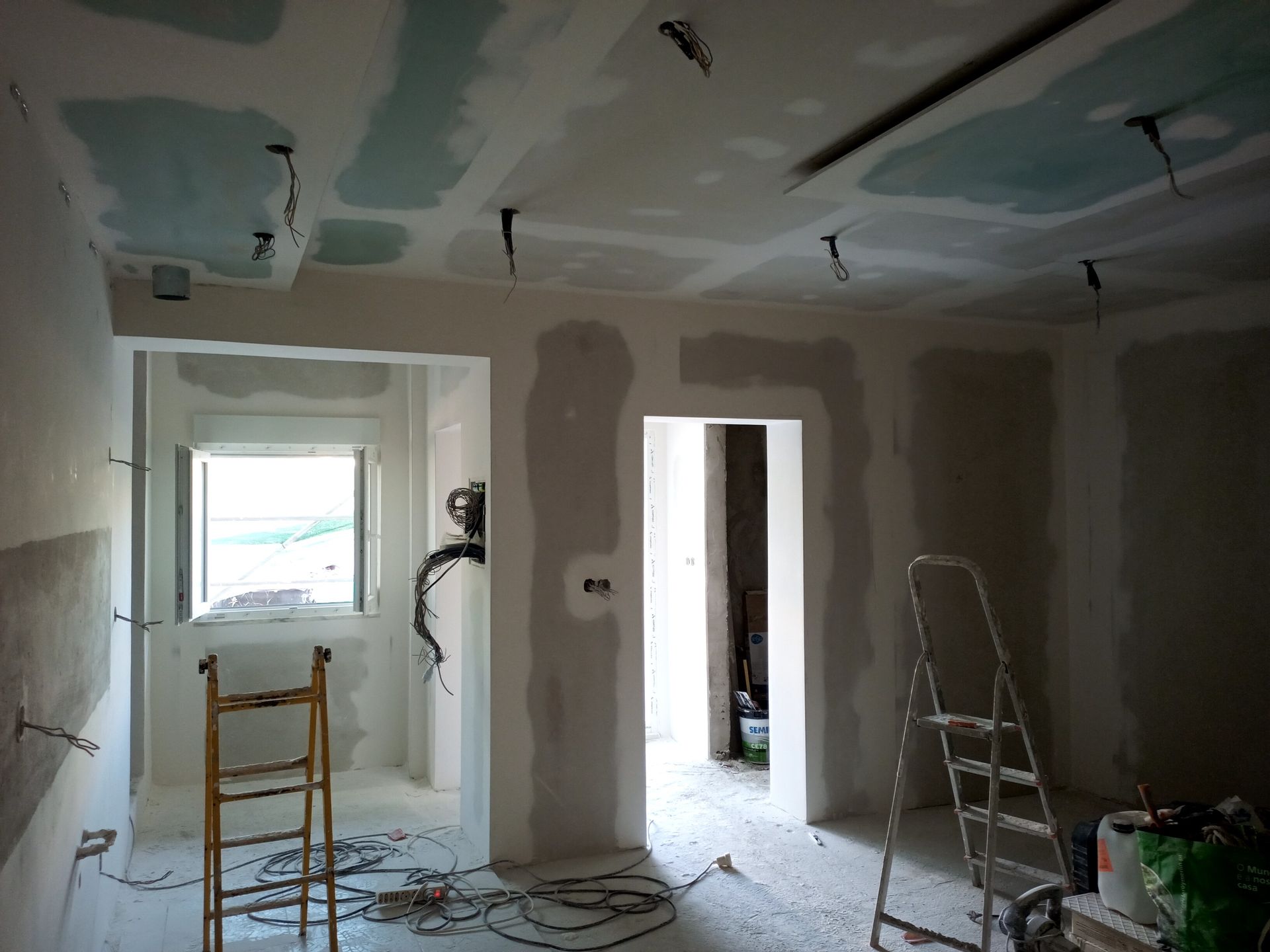 Quarto interno em construção, com drywall instalado nas paredes e no teto, e fiação elétrica exposta. Há uma escada disponível.