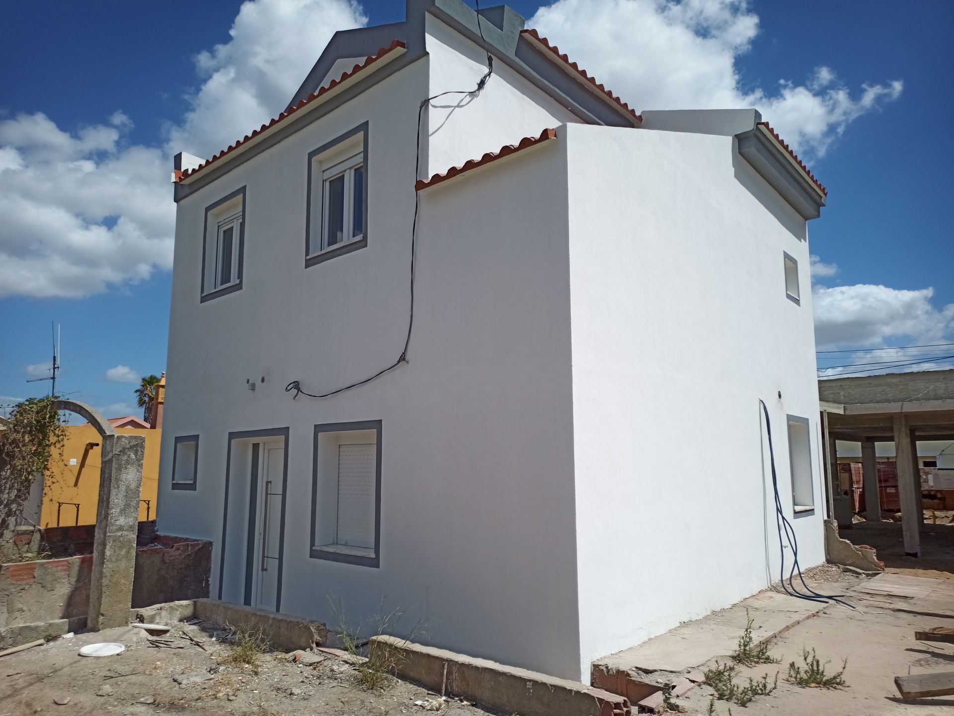 Casa branca de dois andares com detalhes em cinza contra um céu azul com nuvens. Entulho de construção em primeiro plano.