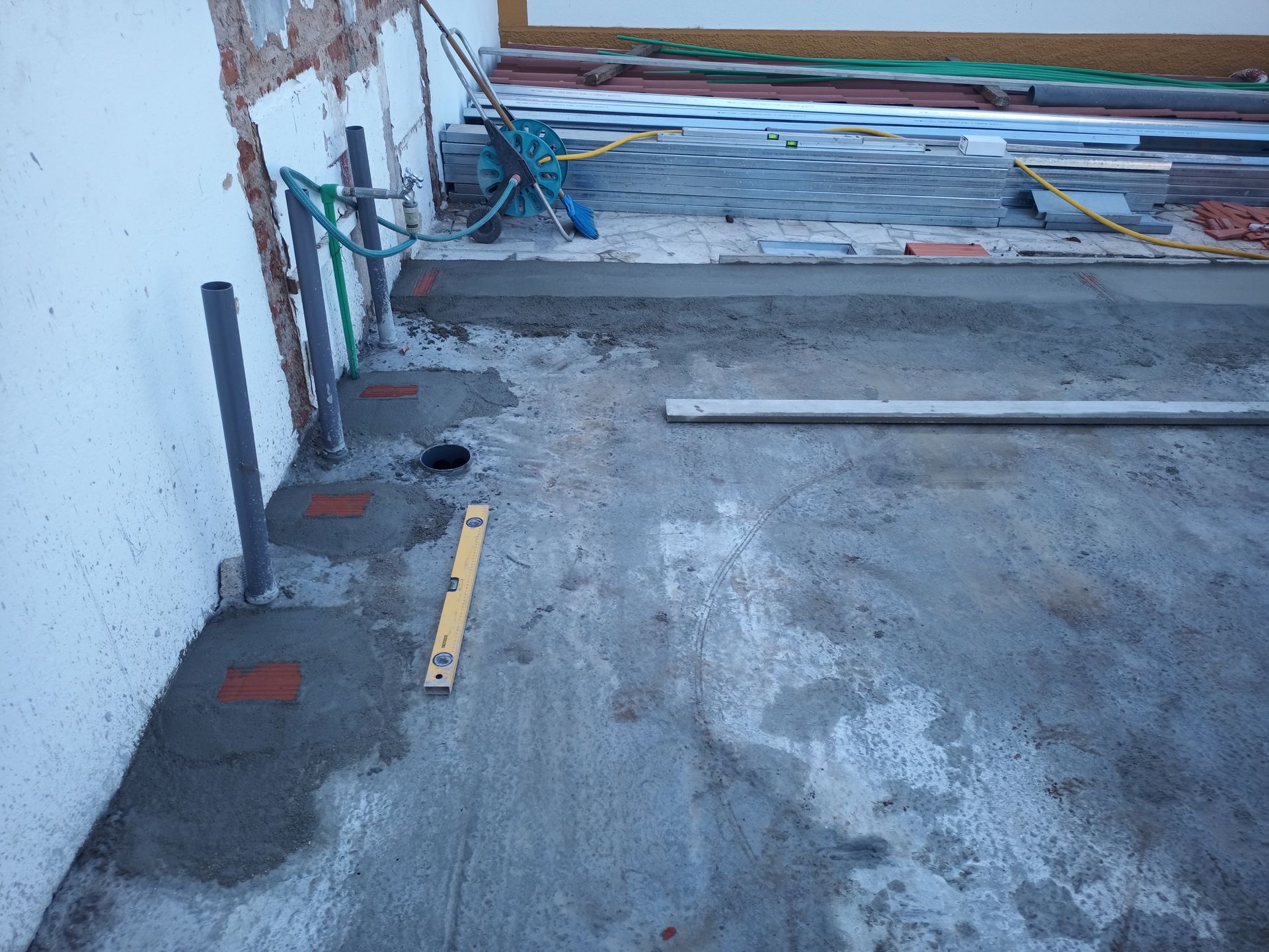 Canteiro de obras: Piso de concreto com tubulações hidráulicas, nível e materiais de construção encostados na parede.