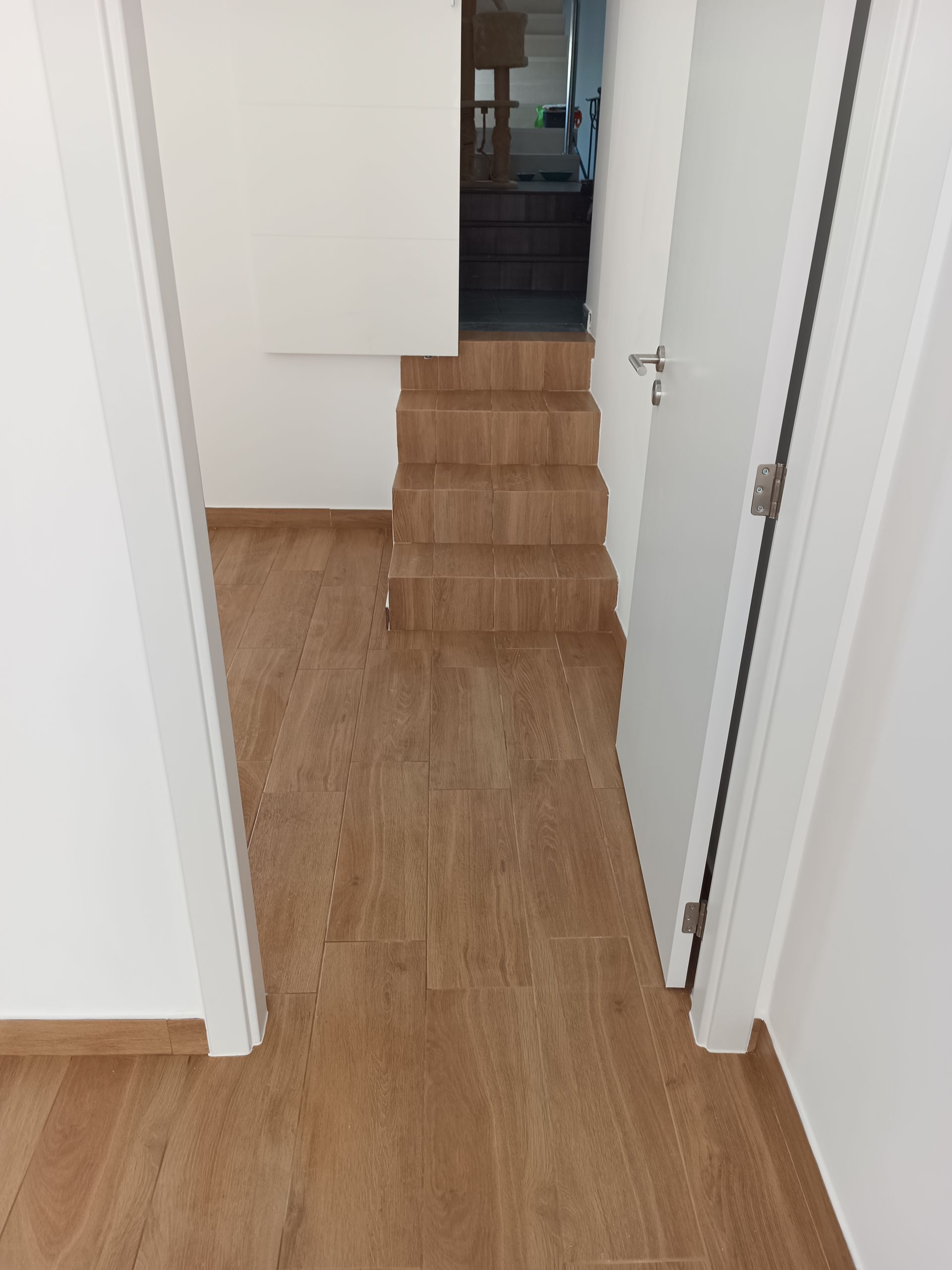 Corredor com piso de madeira que leva à escada; paredes brancas, porta fechada à direita, porta à esquerda.