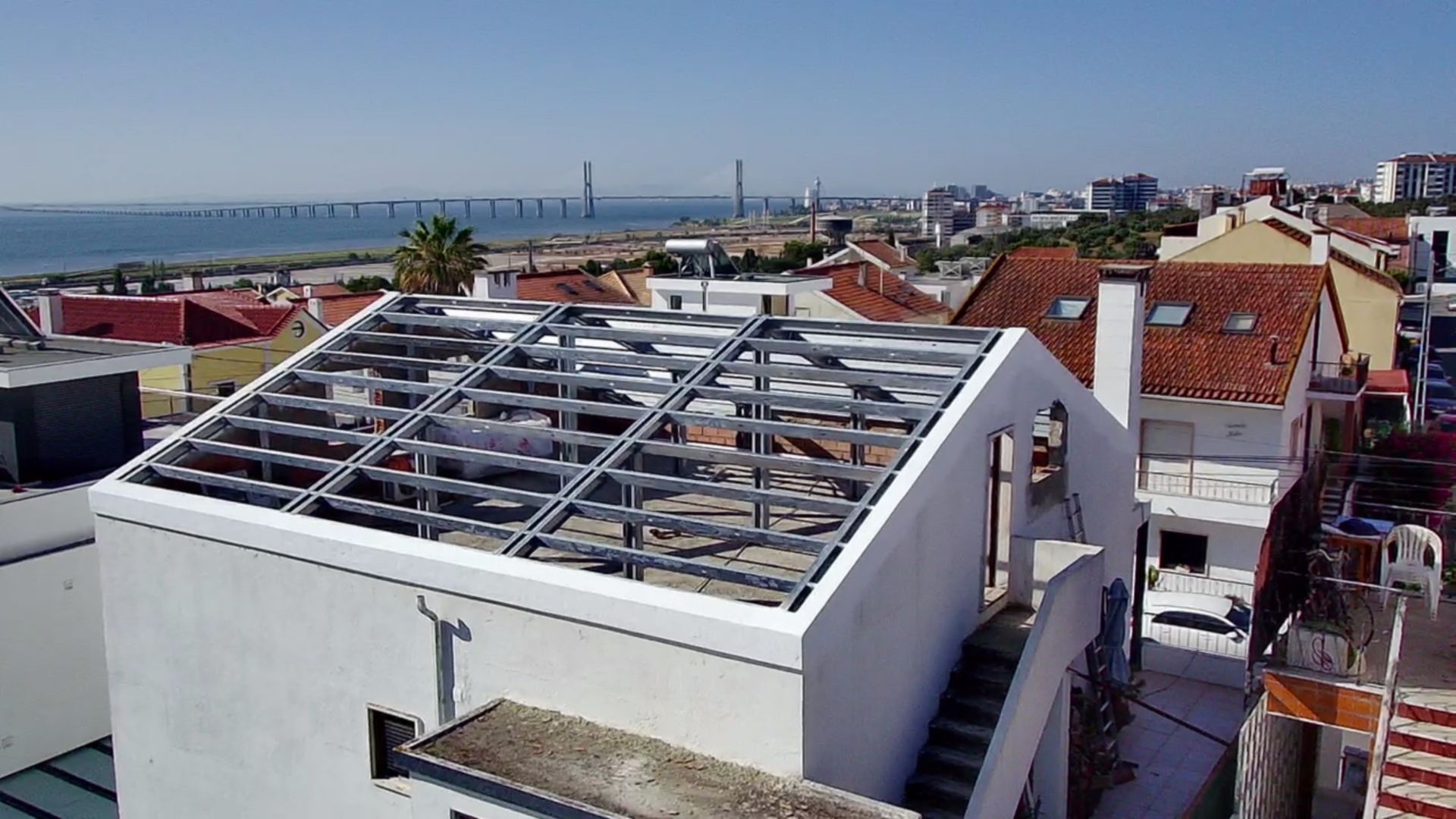 Prédio branco com cobertura de estrutura metálica em construção, paisagem urbana e ponte ao fundo.
