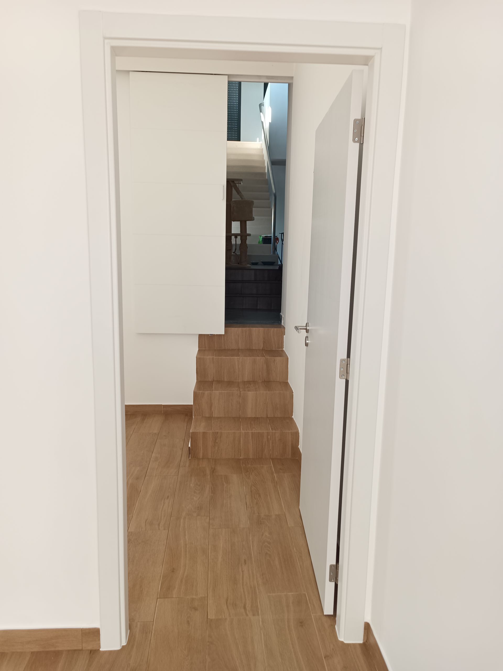 A porta aberta revela uma escada interna e uma porta parcialmente aberta, tudo em branco com piso de madeira.