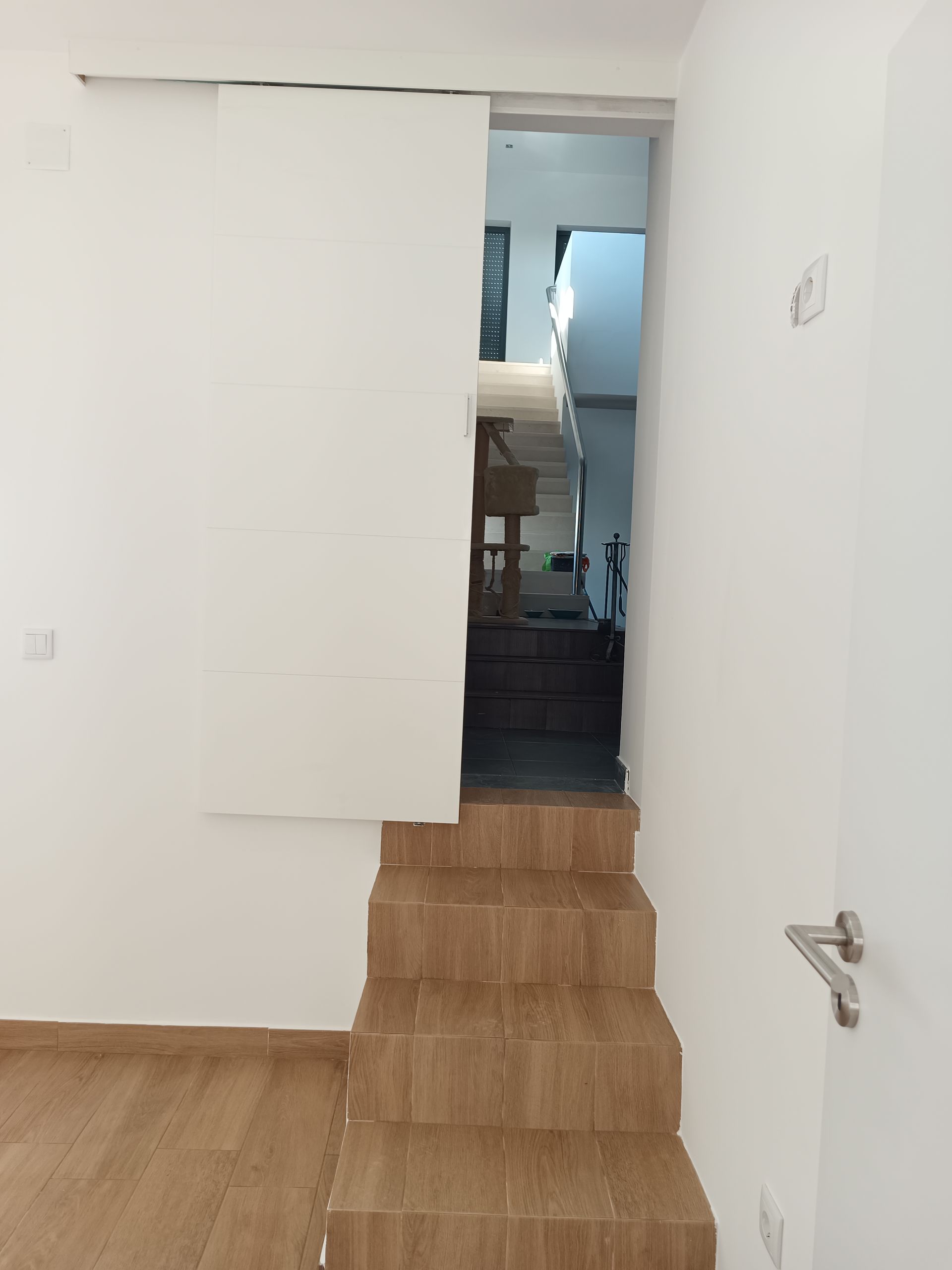 Escada que leva a um nível superior. Degraus de madeira, paredes brancas, porta entreaberta e um vislumbre do interior.