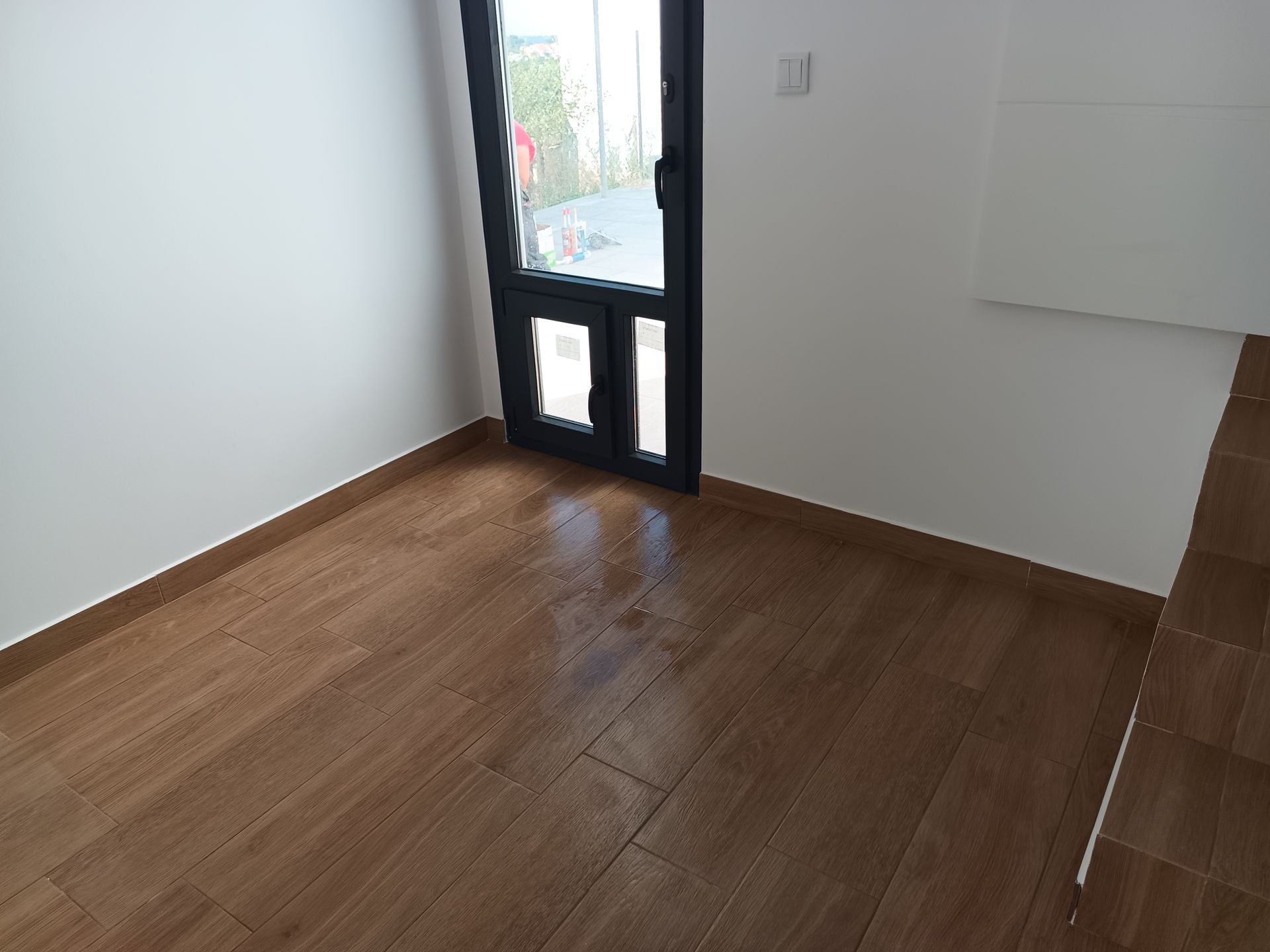 Quarto vazio com piso de madeira, porta com moldura preta e paredes brancas.