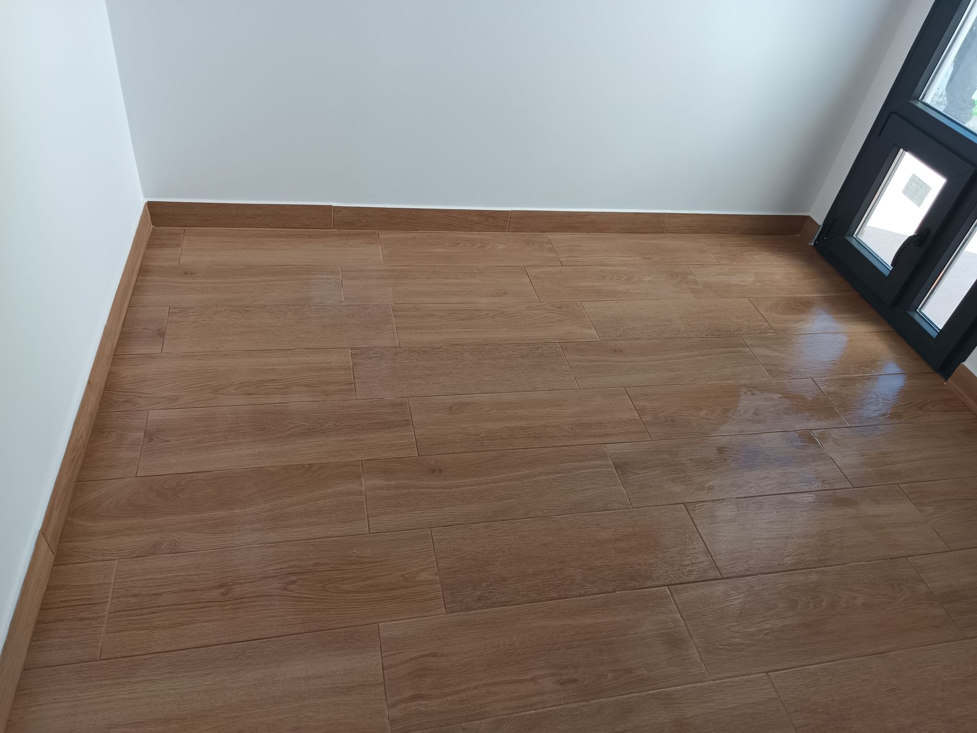 Piso de azulejo marrom em um cômodo com paredes brancas, rodapé e janela com moldura preta.