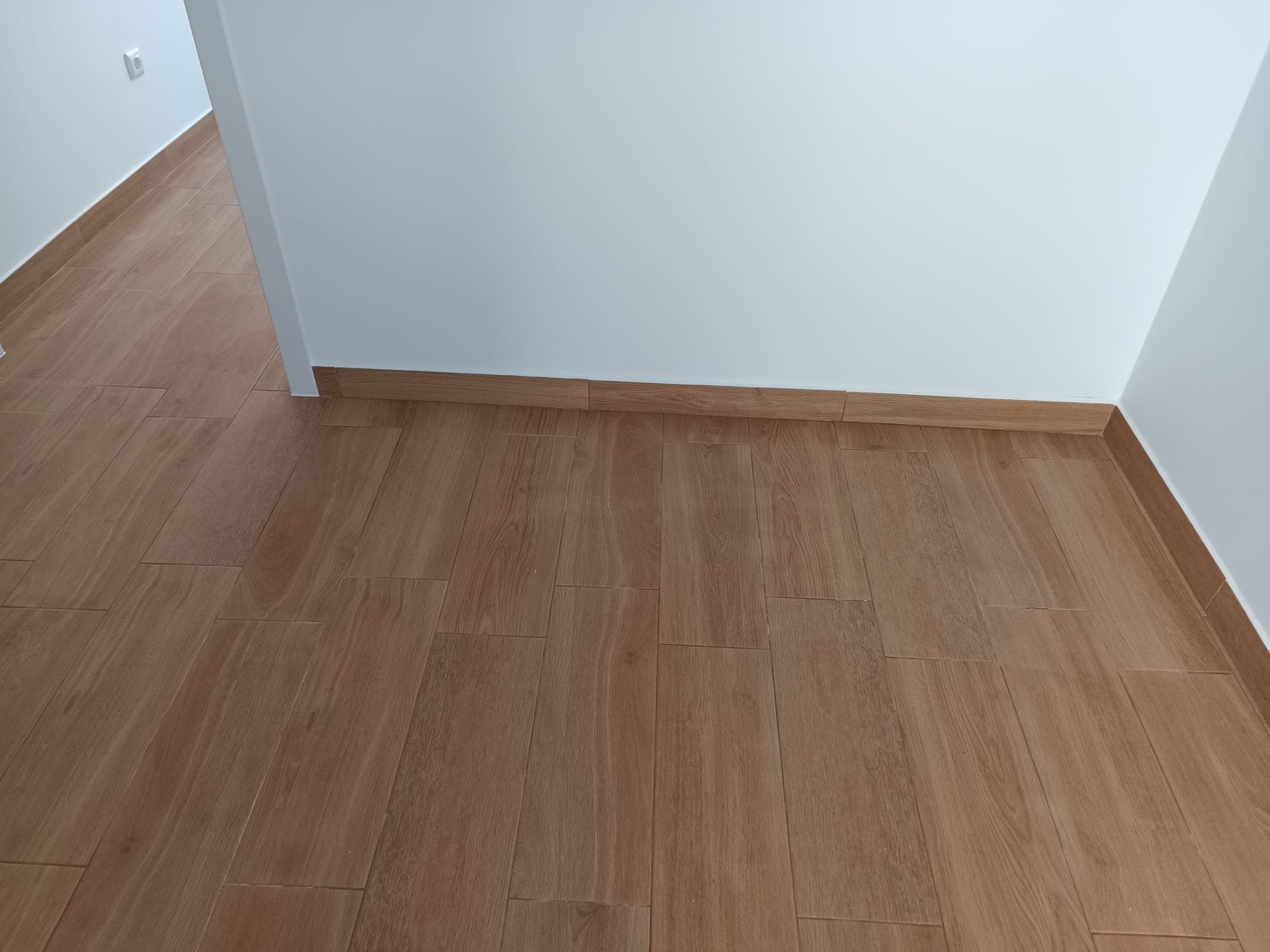 Piso com aparência de madeira, com tábuas marrom-claras e paredes brancas; rodapés marrons revestem as paredes.