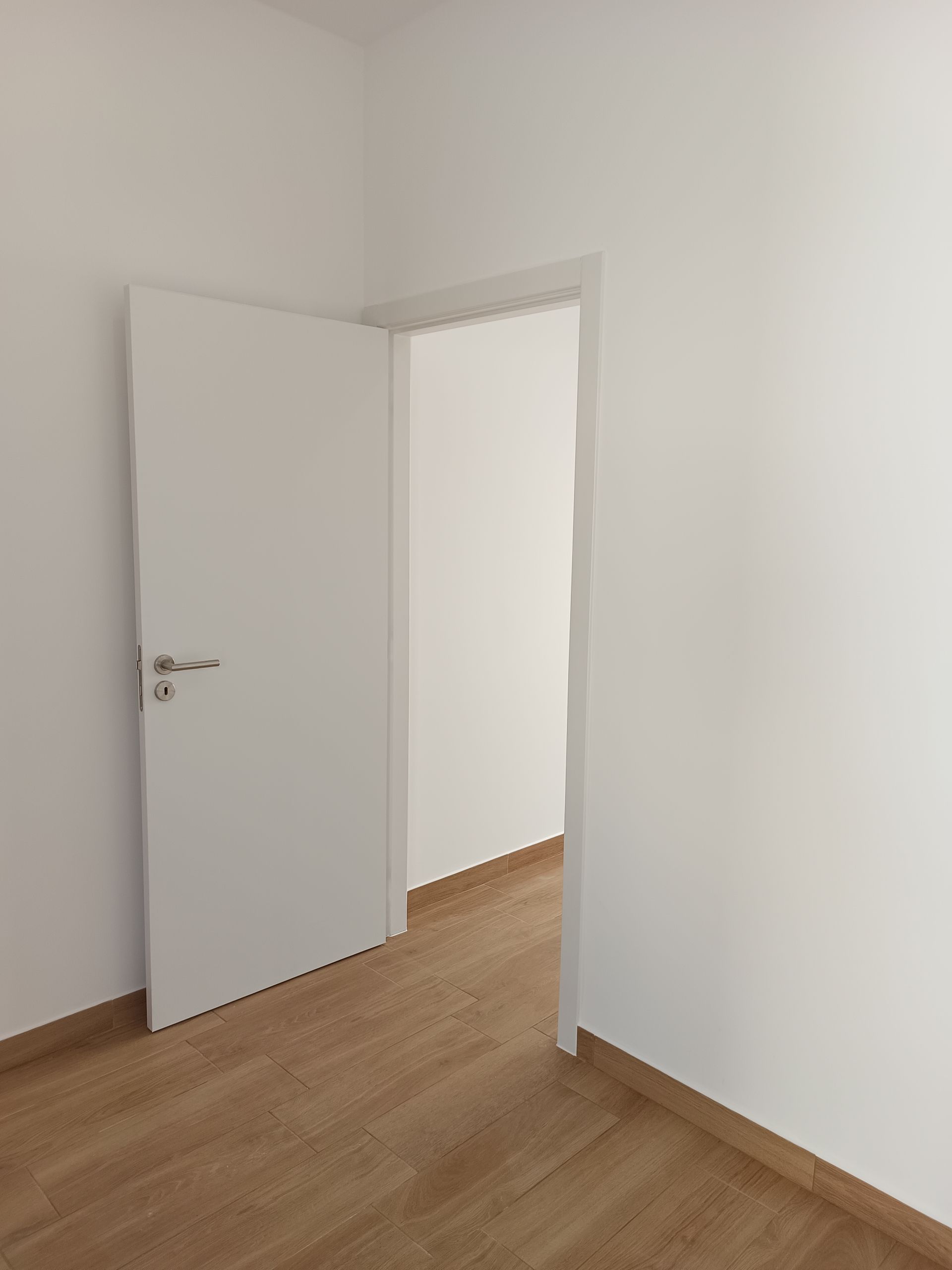 Porta branca entreaberta em um quarto com piso de madeira clara e paredes brancas.