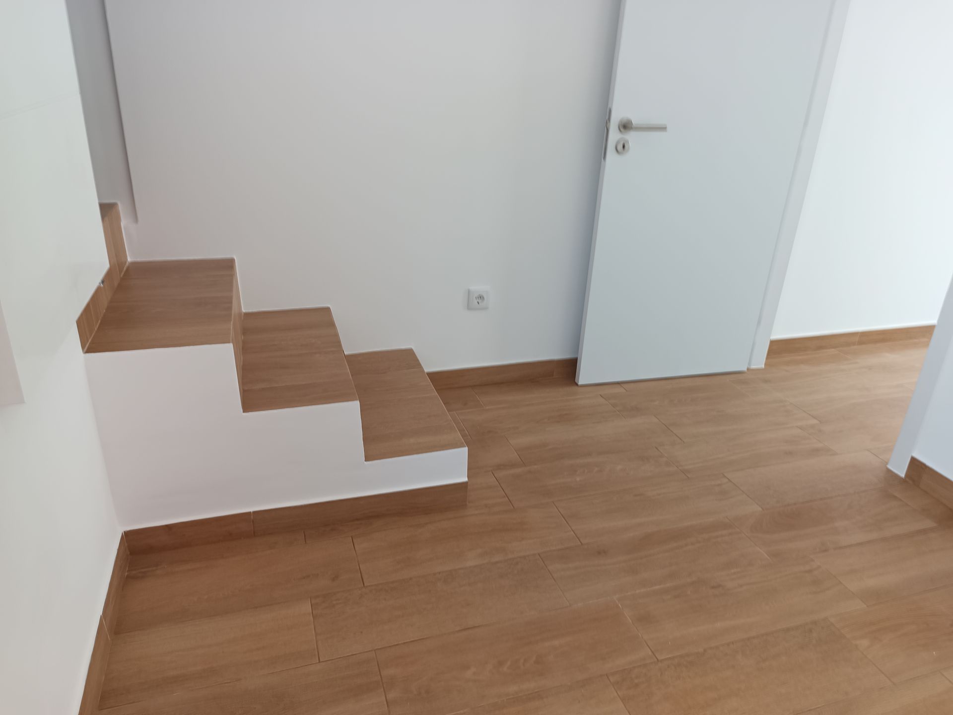 Pequena escada com três degraus, piso de ladrilho de madeira clara, paredes e porta brancas.