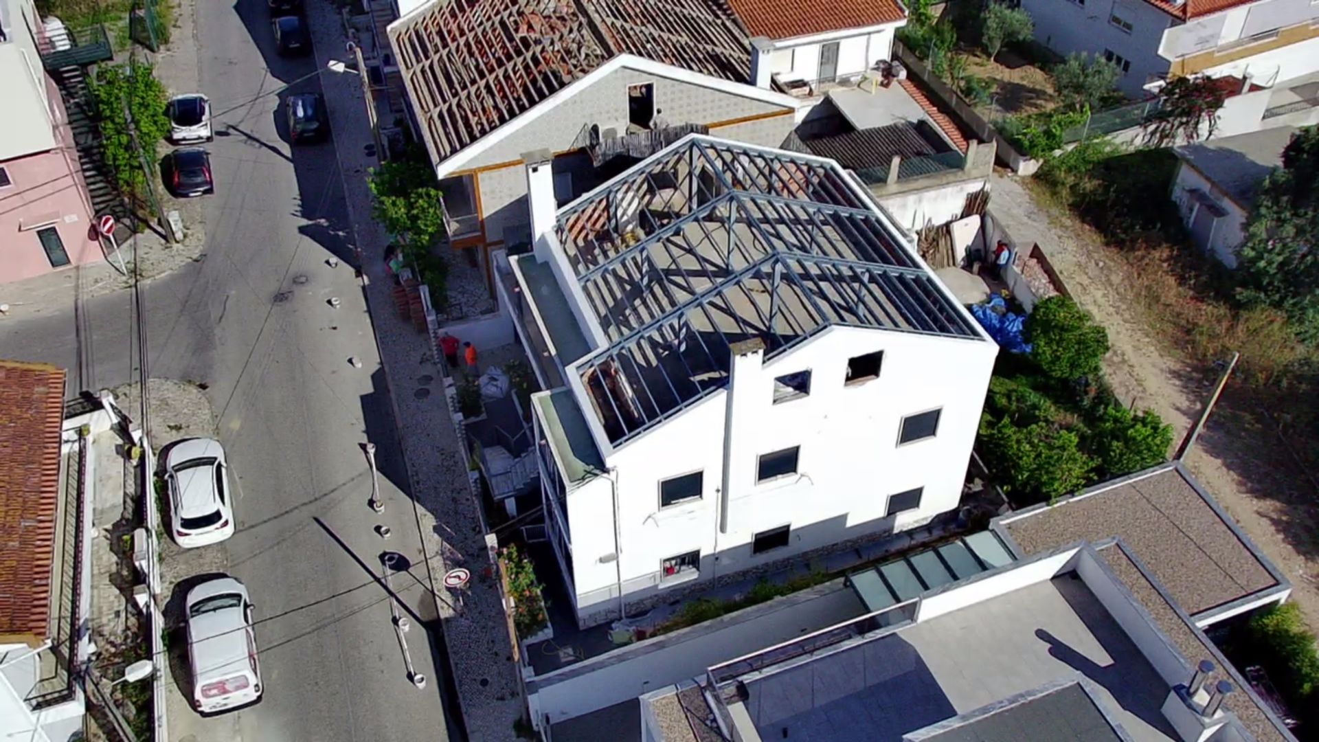 Vista aérea de um edifício branco em construção, com o telhado parcialmente exposto e a rua em frente.