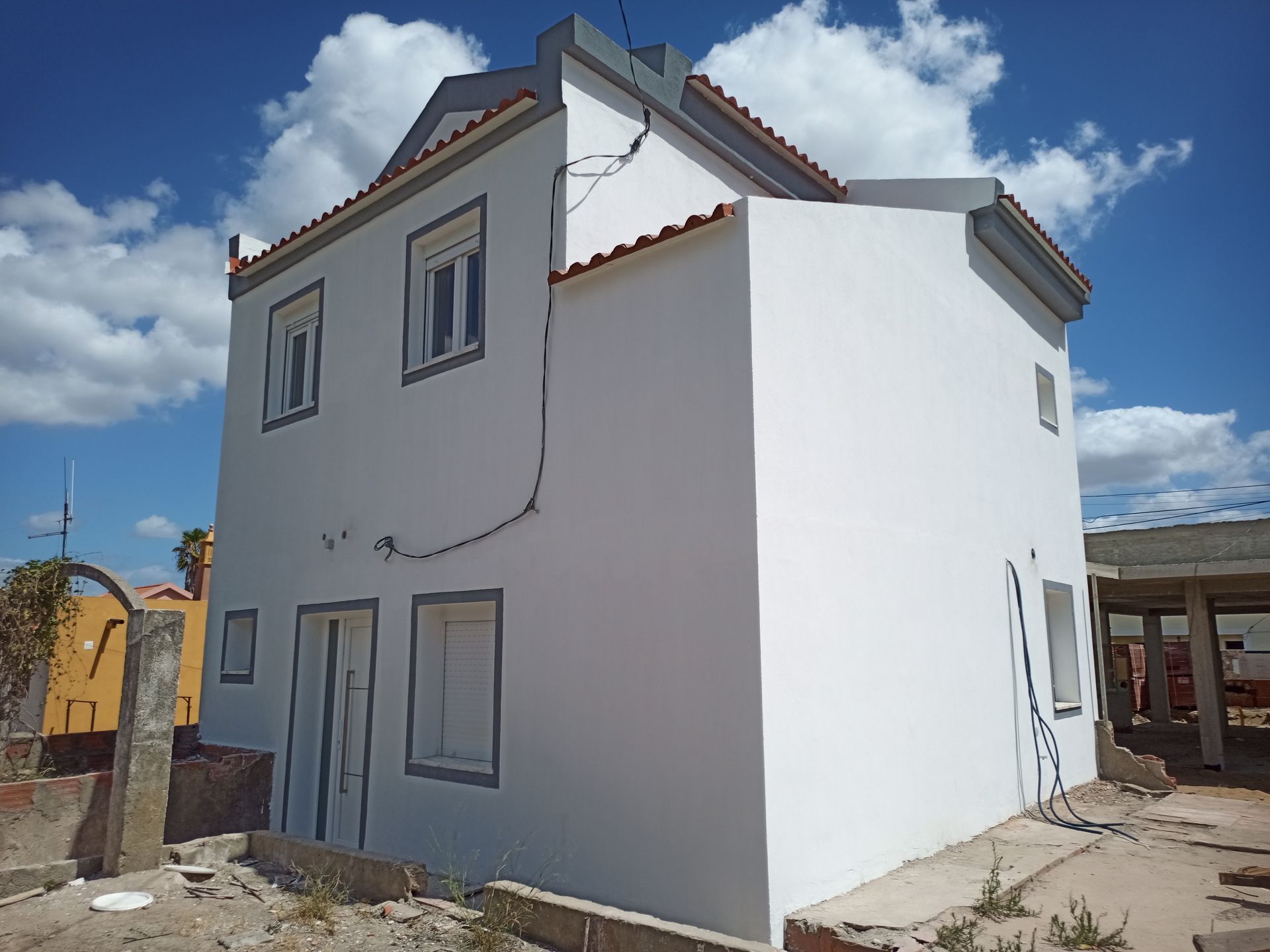 Prédio branco de dois andares com detalhes em cinza e rachaduras visíveis, contra um céu azul com nuvens.