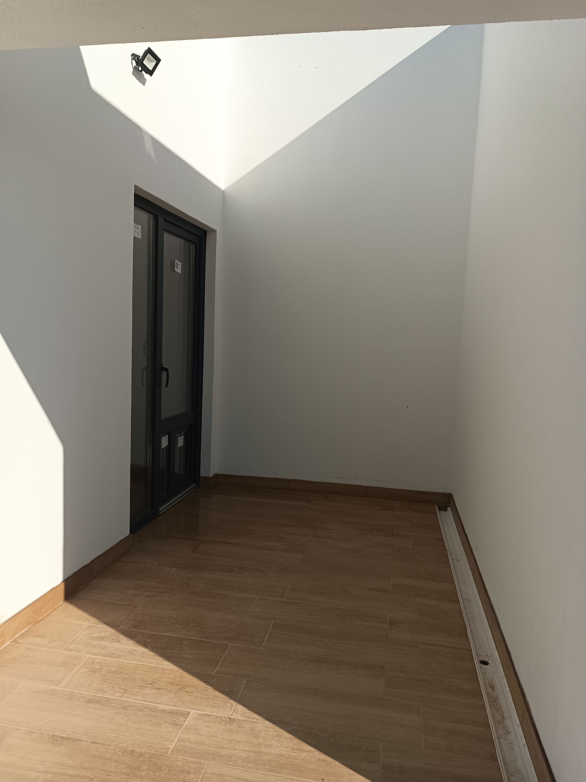 Área externa fechada com paredes brancas, piso de azulejo e porta de correr com moldura escura. A luz do sol projeta sombras.