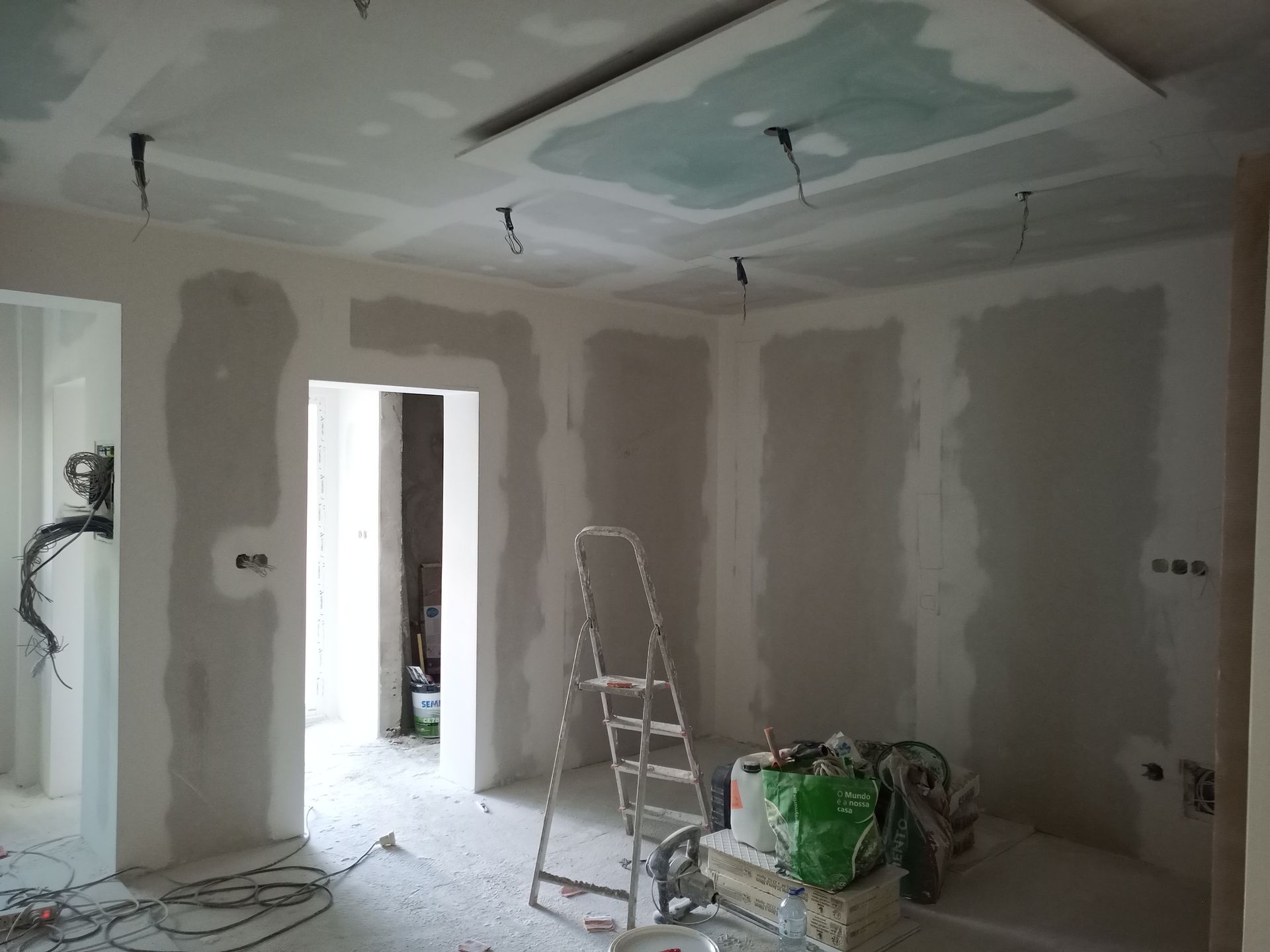 Quarto interno com instalação de drywall; teto inacabado com painel embutido e fiação suspensa; escada de mão.