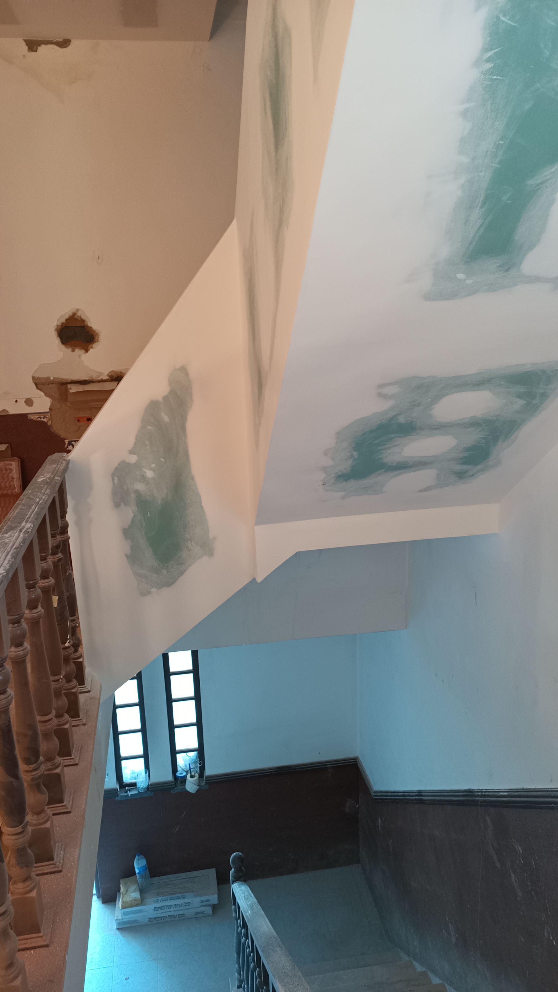 Escada com tinta descascando, apresentando manchas verdes no teto e nas paredes. Corrimão de madeira e janela visíveis.