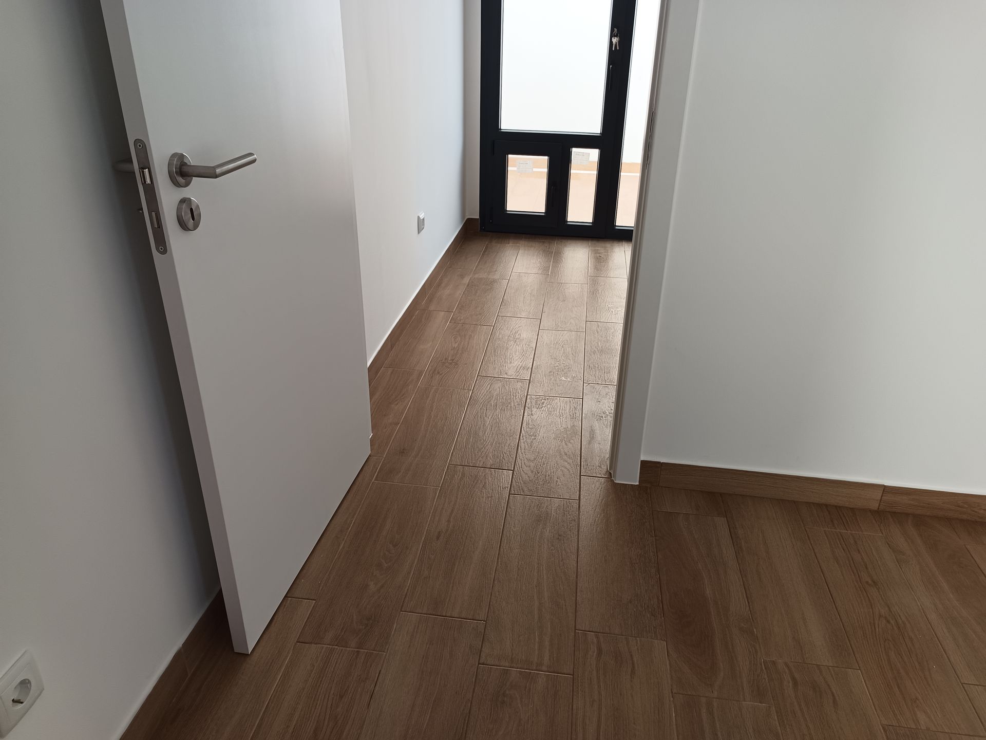 Corredor com piso marrom claro, paredes brancas e uma porta que dá acesso a um cômodo com porta e janela pretas.