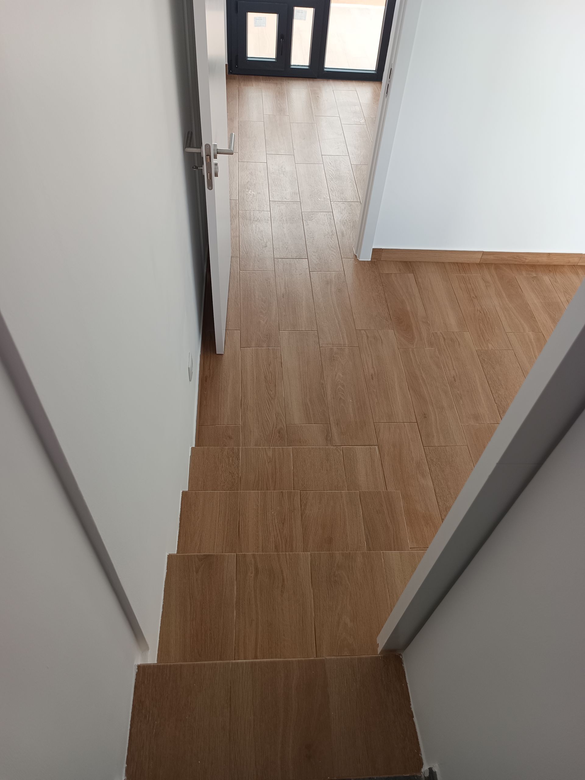 Escada com degraus e piso de madeira, que leva a uma porta e janela no final de um corredor.