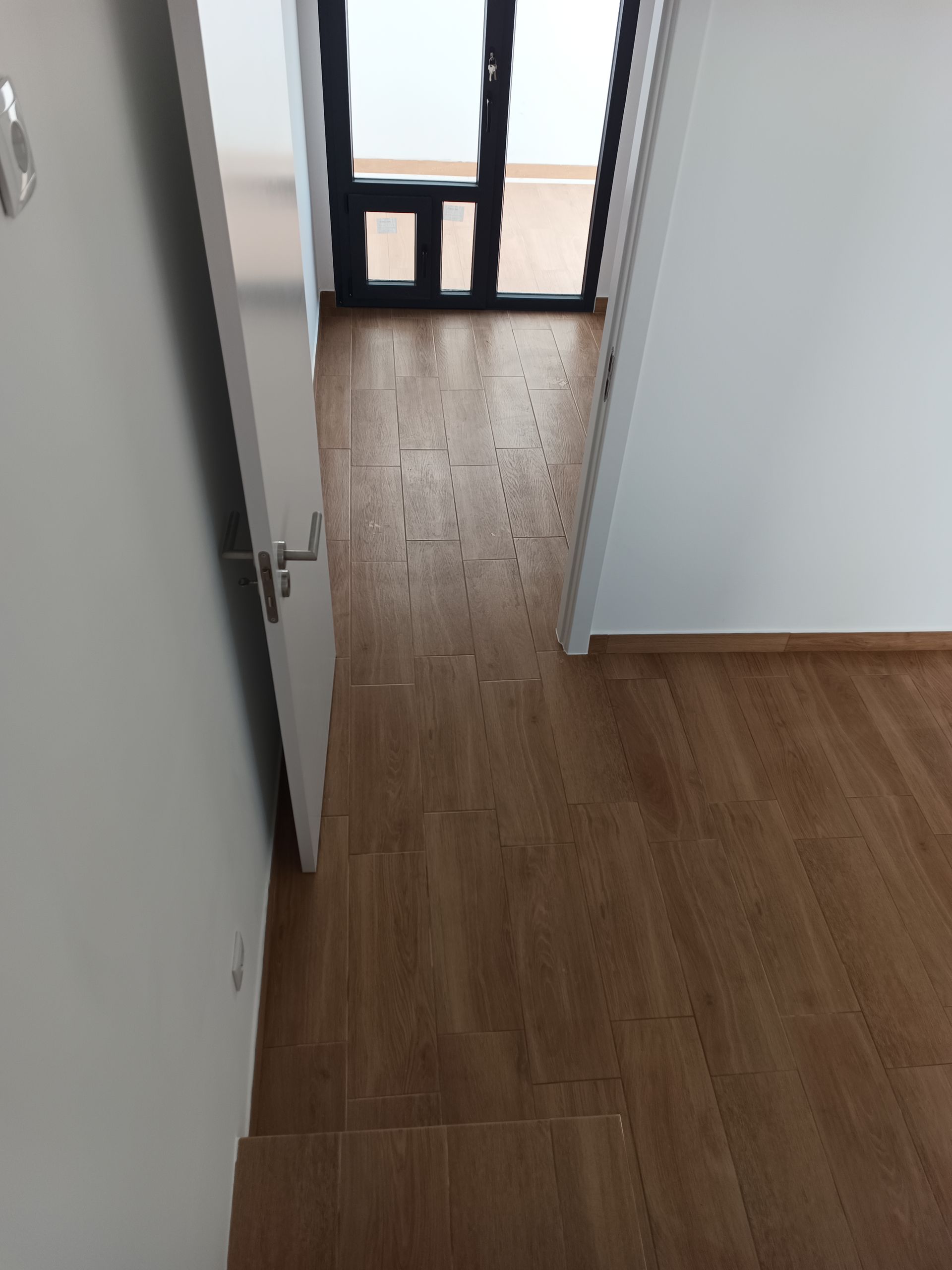 Porta de entrada que dá acesso a um quarto com piso de azulejo imitando madeira e uma porta de vidro para uma varanda.