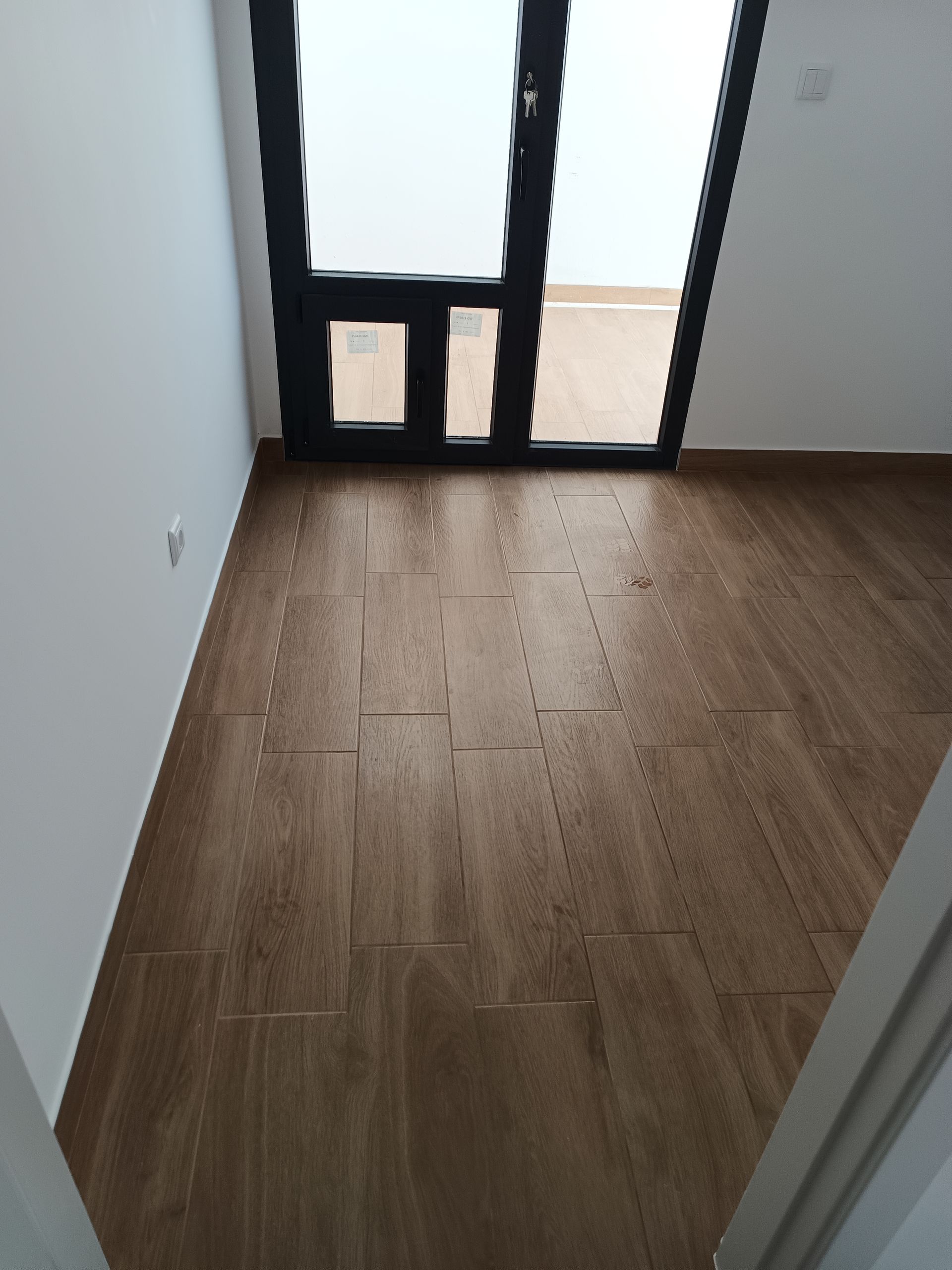 Piso imitando madeira marrom em um cômodo com porta de moldura preta que dá para uma área externa. Paredes brancas.