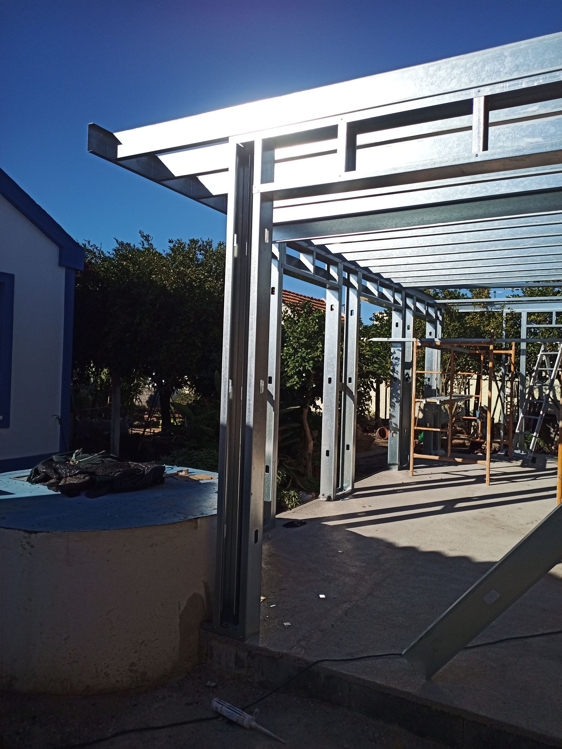 Estrutura de aço de um pátio com céu azul ao fundo e uma casa.