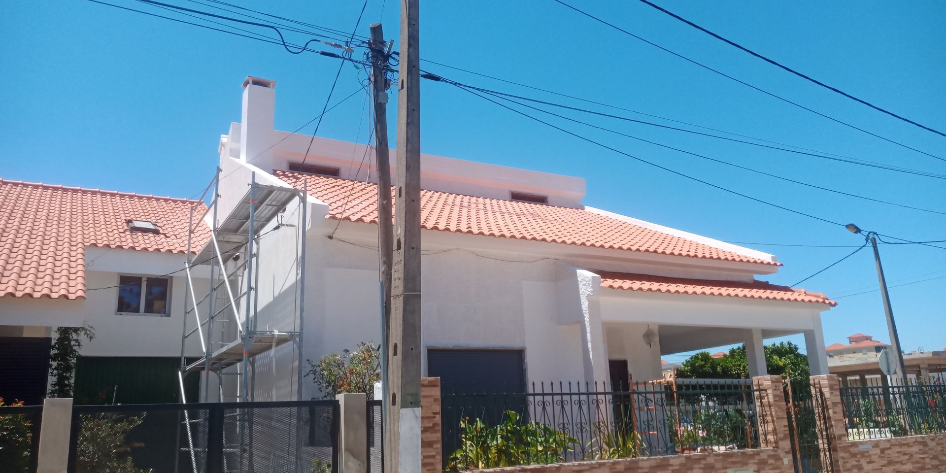 Casa branca com telhado de telha laranja, portão preto, andaimes e poste telefônico contra um céu azul.