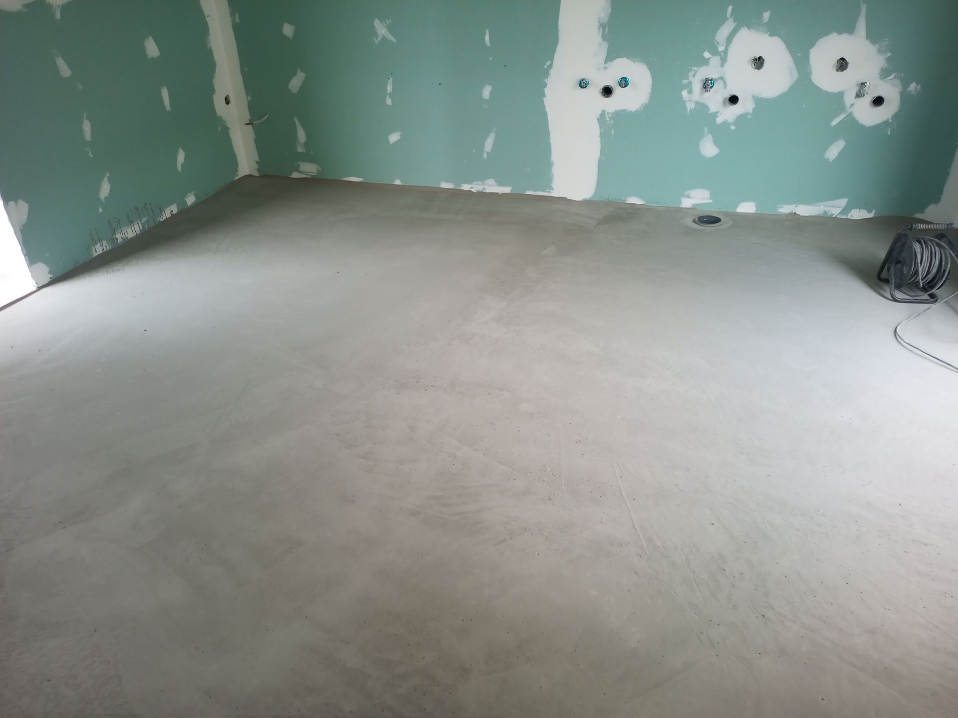 Sala interna com piso de concreto cinza alisado e paredes de gesso cartonado azul-esverdeado sem acabamento.