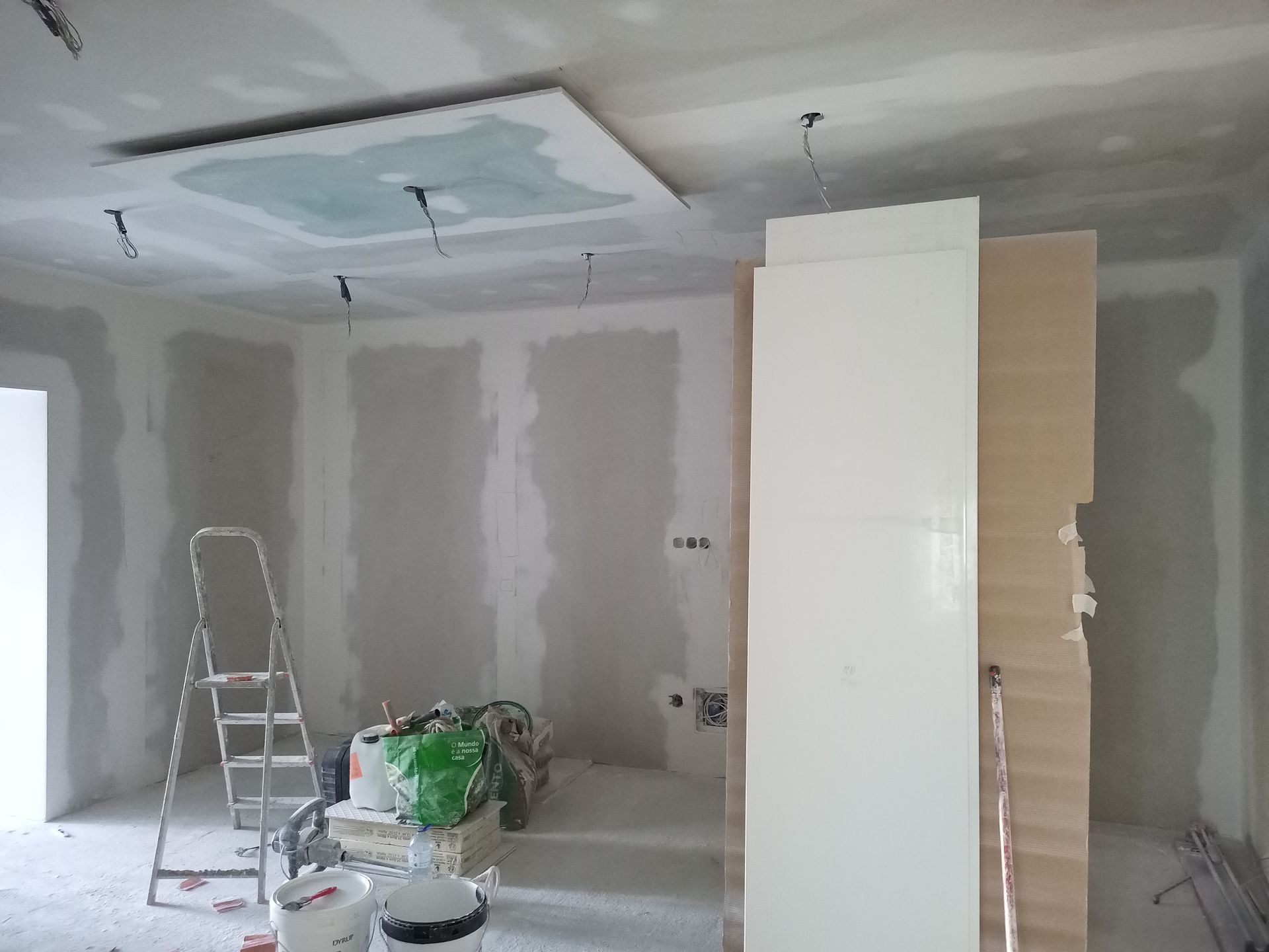 Sala em construção com drywall, teto rebaixado e armários; uma escada e baldes são visíveis.