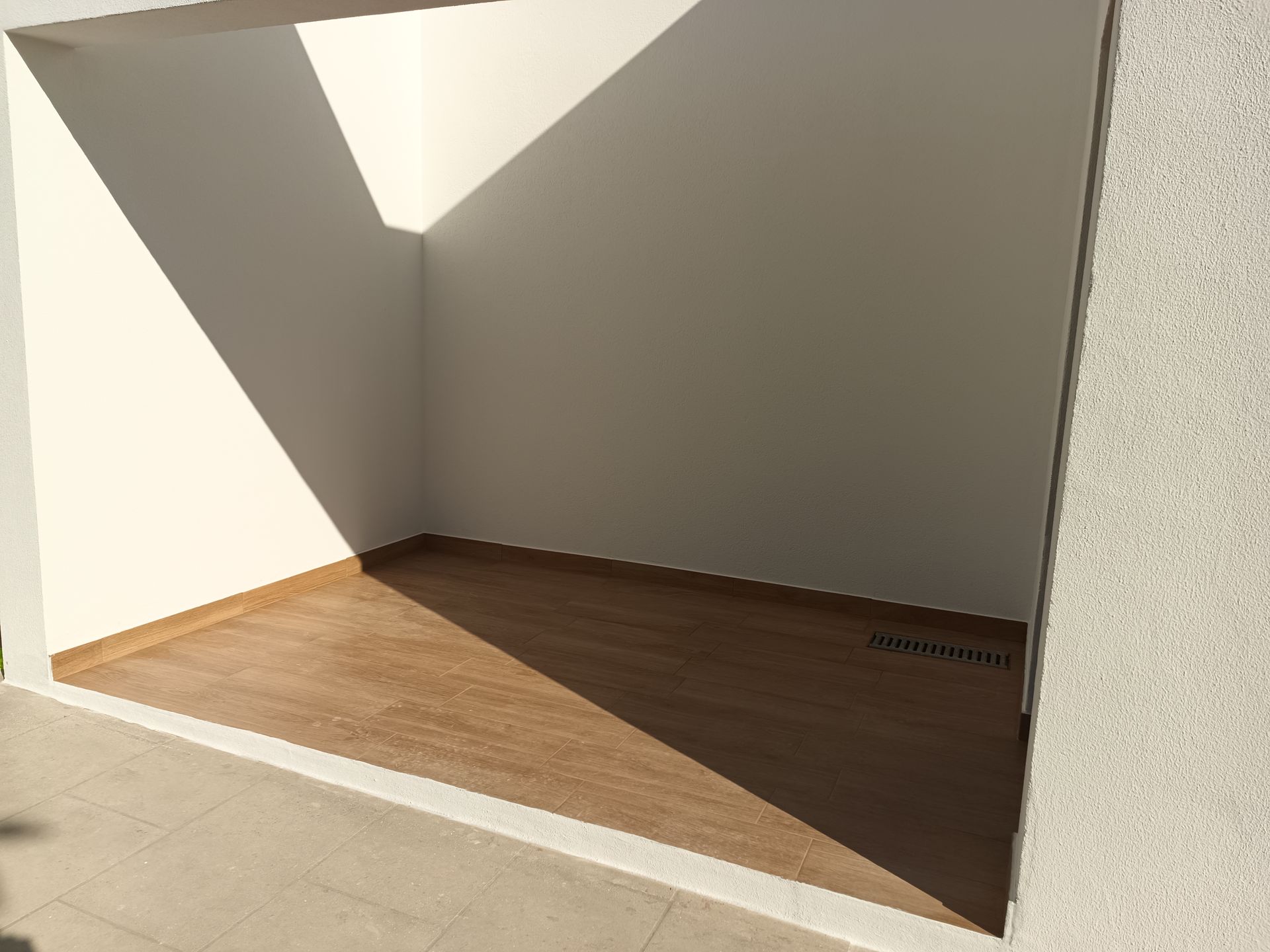 Chuveiro externo vazio com piso que imita madeira e paredes brancas. A luz do sol cria sombras.