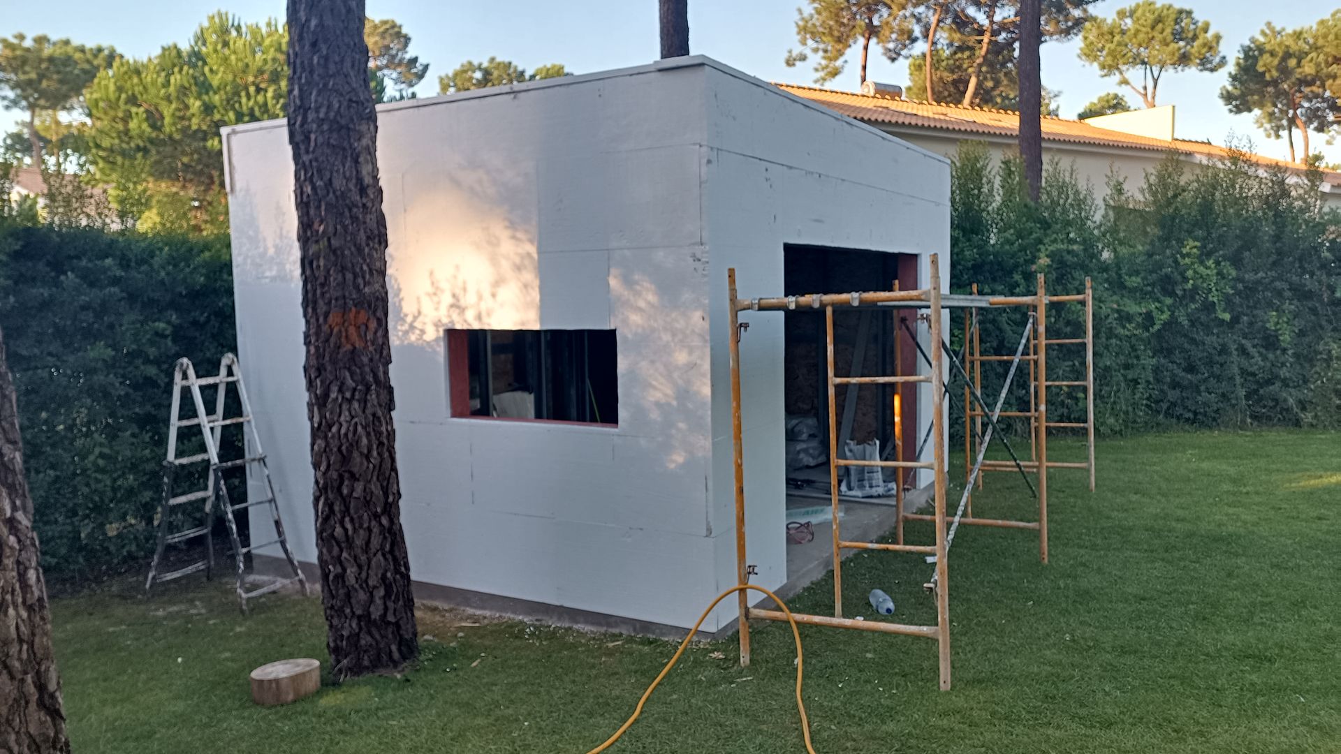 Construção de um prédio branco com andaimes, duas escadas e uma árvore em um gramado verde.