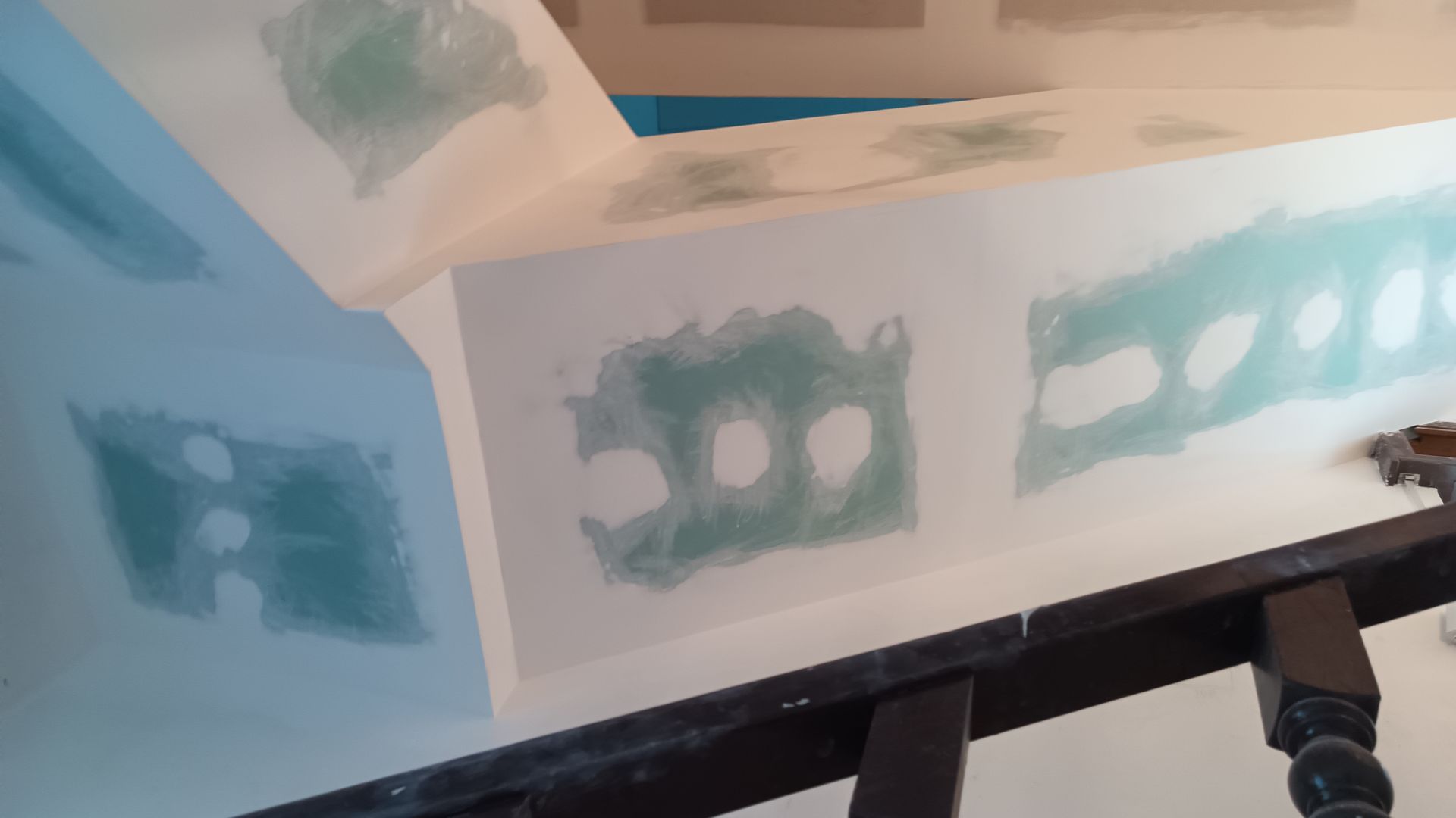 Reparo de drywall em parede e teto brancos. Massa de remendo verde é visível. Um corrimão de madeira escura está em primeiro plano.