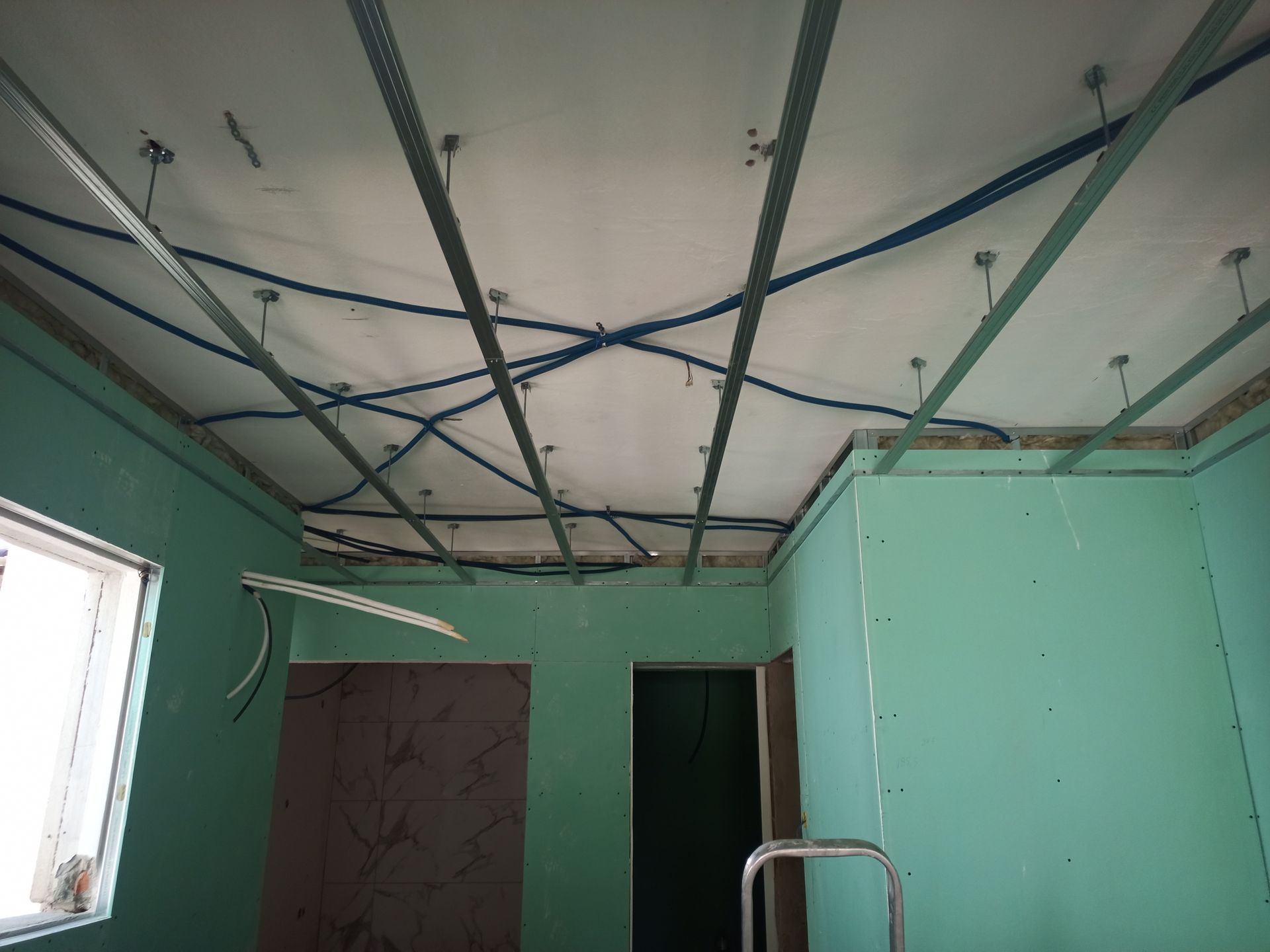 Sala em construção com vigas de sustentação do teto, fiação e drywall expostos. Paredes verdes, janela, porta e escada são visíveis.
