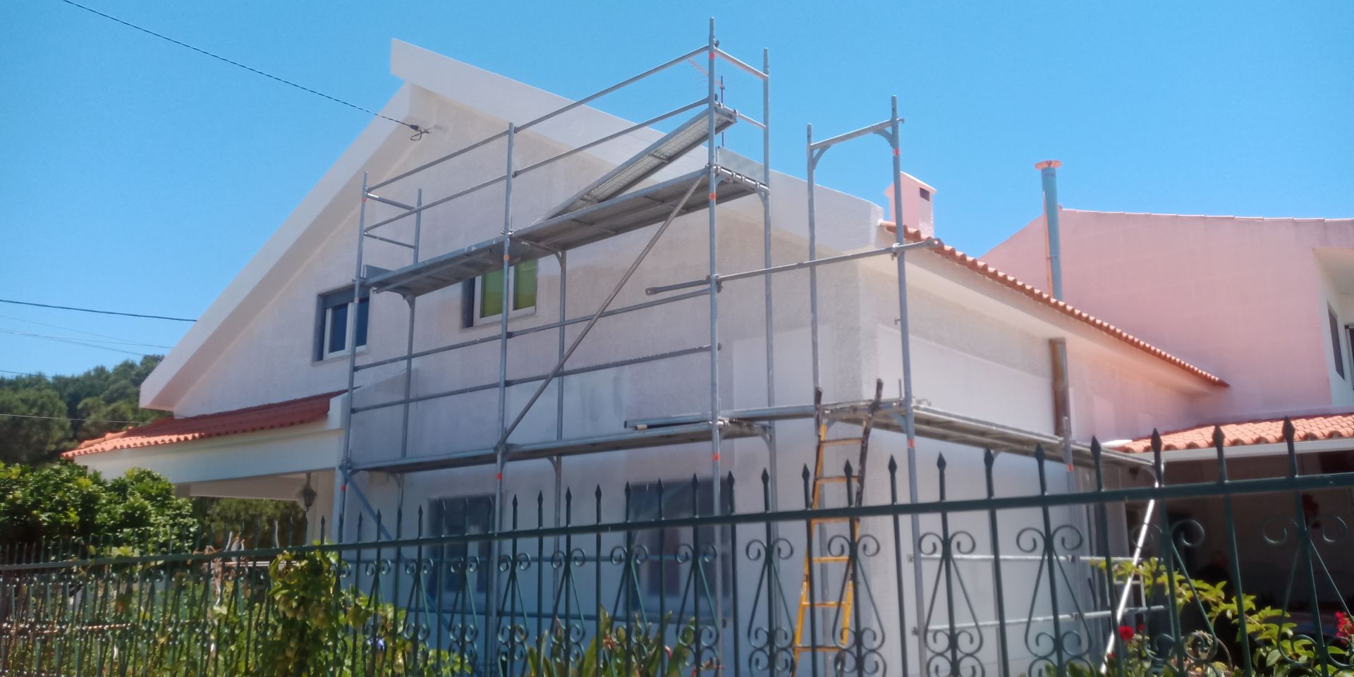 Casa branca com andaimes, em construção num dia ensolarado. Cerca metálica em primeiro plano.